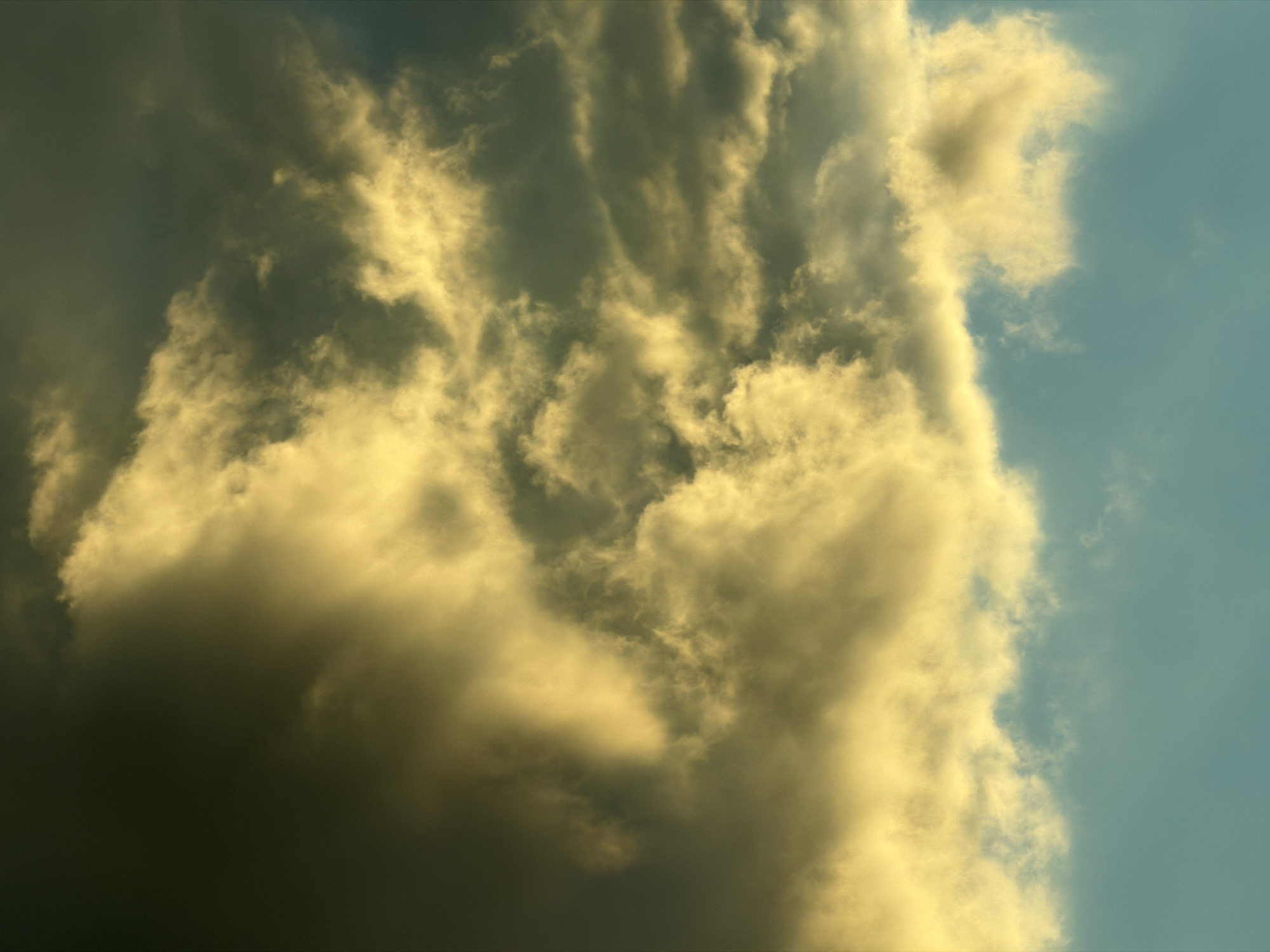 The image is a vertical, eye-level, close-up shot of dramatic, turbulent clouds against a teal-colored sky. The clouds are lit from the left, with bright yellow and golden hues highlighting their fluffy textures, while the right side of the image is darker with deep grays and blacks. A band of lighter, more wispy clouds separates the upper, darker clouds from the clearer sky below. The scene appears to be an outdoor sky view, possibly taken in Studena, Bulgaria, given the context provided. There are no people, subjects, objects, or actions depicted.