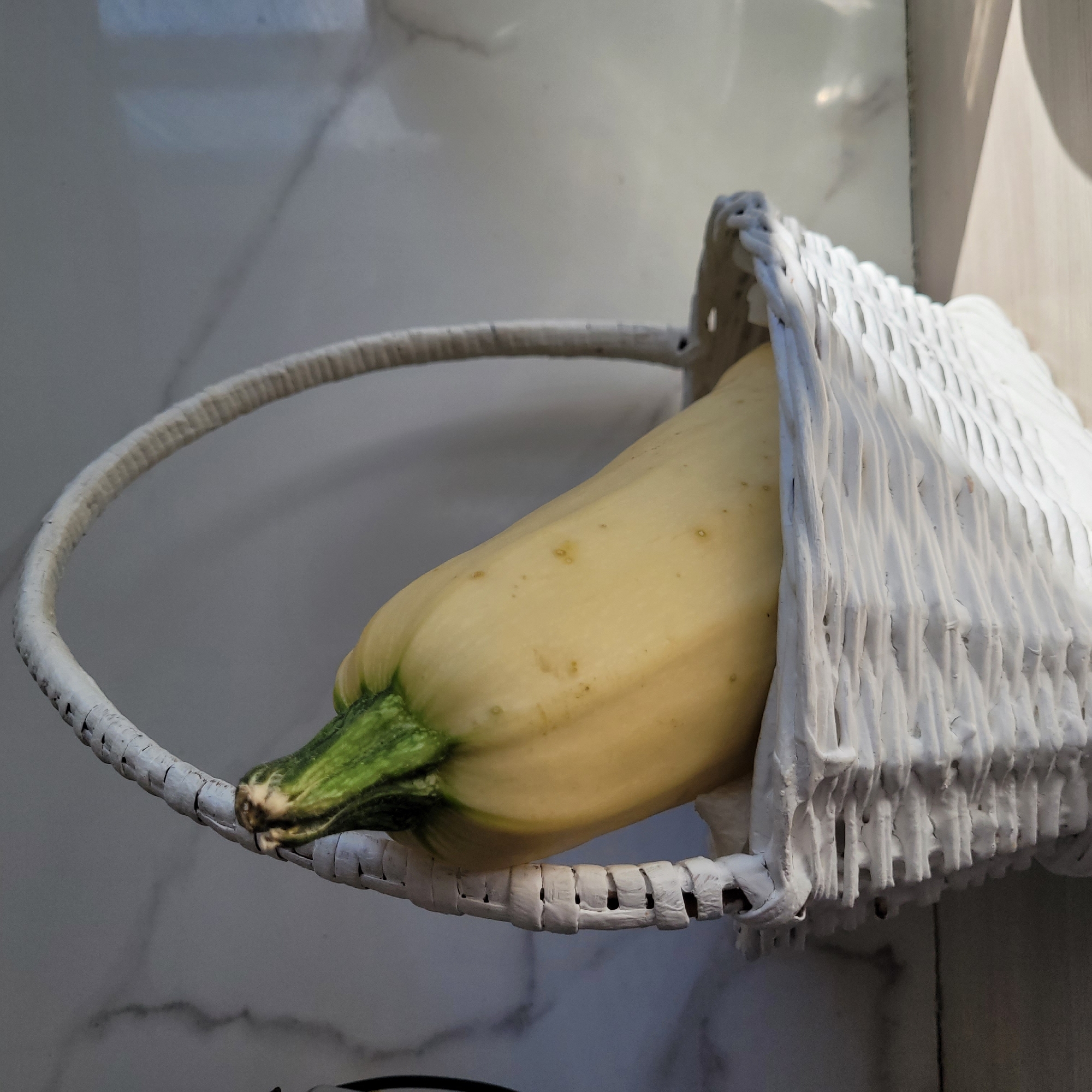 A light yellow zucchini with a green stem rests inside a white woven basket. The basket is placed on a light-colored surface, possibly a countertop or table. In the background, a marbled surface is visible, with shadows cast across it, indicating natural light from the right. The scene appears to be indoors, potentially in a kitchen or dining area. There are no people or animals present. The composition is framed to focus on the zucchini and basket, with the background providing context. The image captures a still life arrangement with a focus on texture and light.