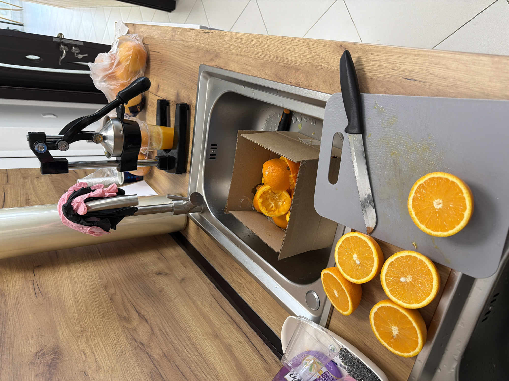 The image shows a kitchen counter with items related to orange juice preparation. A manual citrus juicer is mounted on the left side of the counter. There are oranges in a plastic bag next to the juicer and sliced oranges on a grey cutting board to the right. A knife is also present on the cutting board. In the sink, there is a small cardboard box containing orange peels. A roll of plastic wrap and a pair of pink and black gloves are on the counter in front of the juicer. A plastic container with cleaning supplies is situated near the edge of the counter near the sliced oranges. The countertop has a wood grain finish. It is impossible to confirm the location in the city of Gabrovo, Bulgaria, based on the visual information provided.