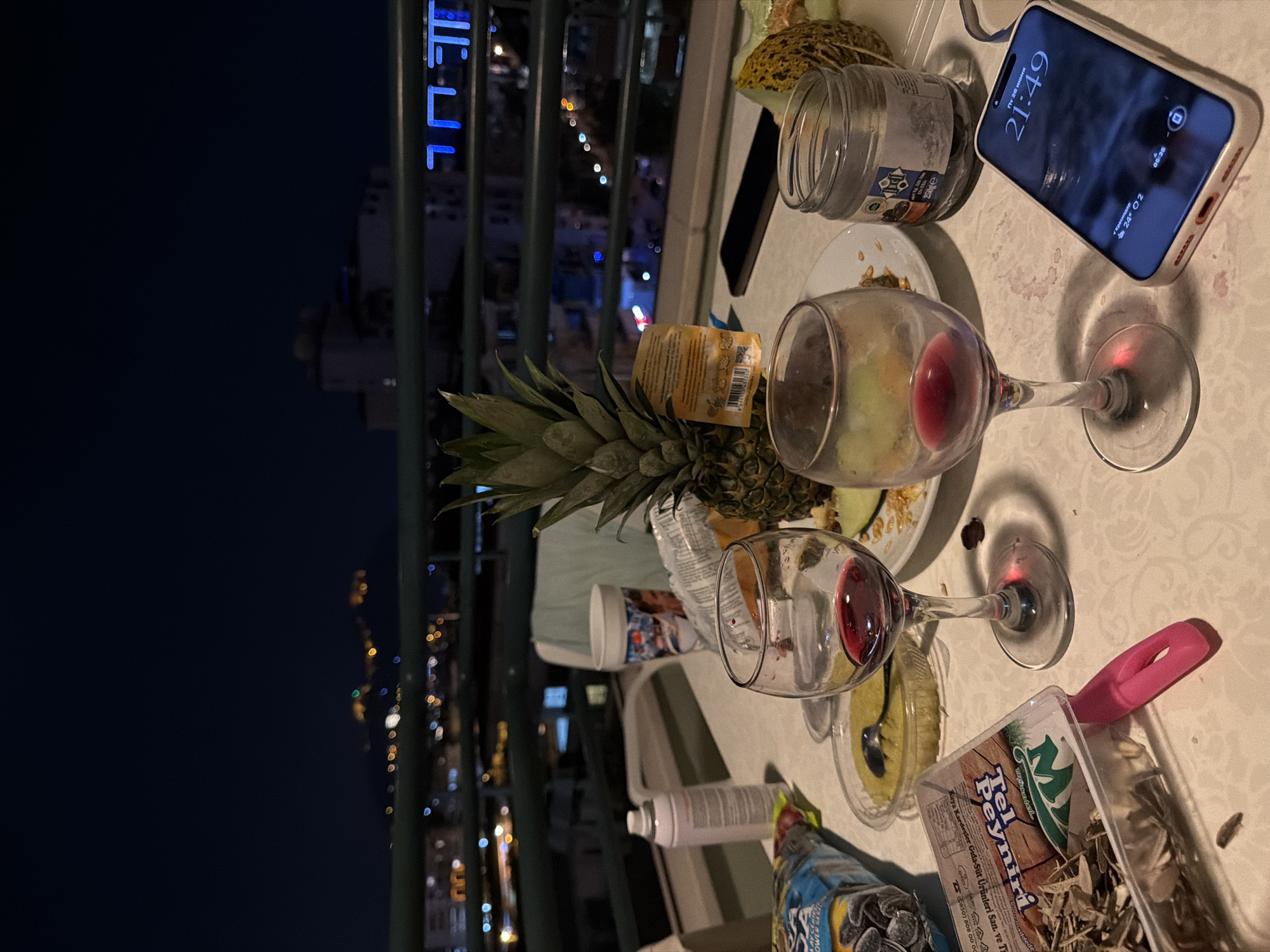 This is a top-down, rotated shot of a messy table on a balcony at night. In the foreground, two wine glasses, one partially filled with red wine, sit on a white tablecloth. A pineapple, a half-eaten melon, and a jar of what appears to be jam or preserves are also on the table, along with a discarded phone displaying the time "21:49" and the date "Fri, June 20". A container of cheese and a bag of sunflower seeds are also visible. In the background, the lights of a city twinkle below, suggesting an urban setting at night. The overall scene conveys a sense of a casual, possibly late-night gathering or a meal that has just concluded.