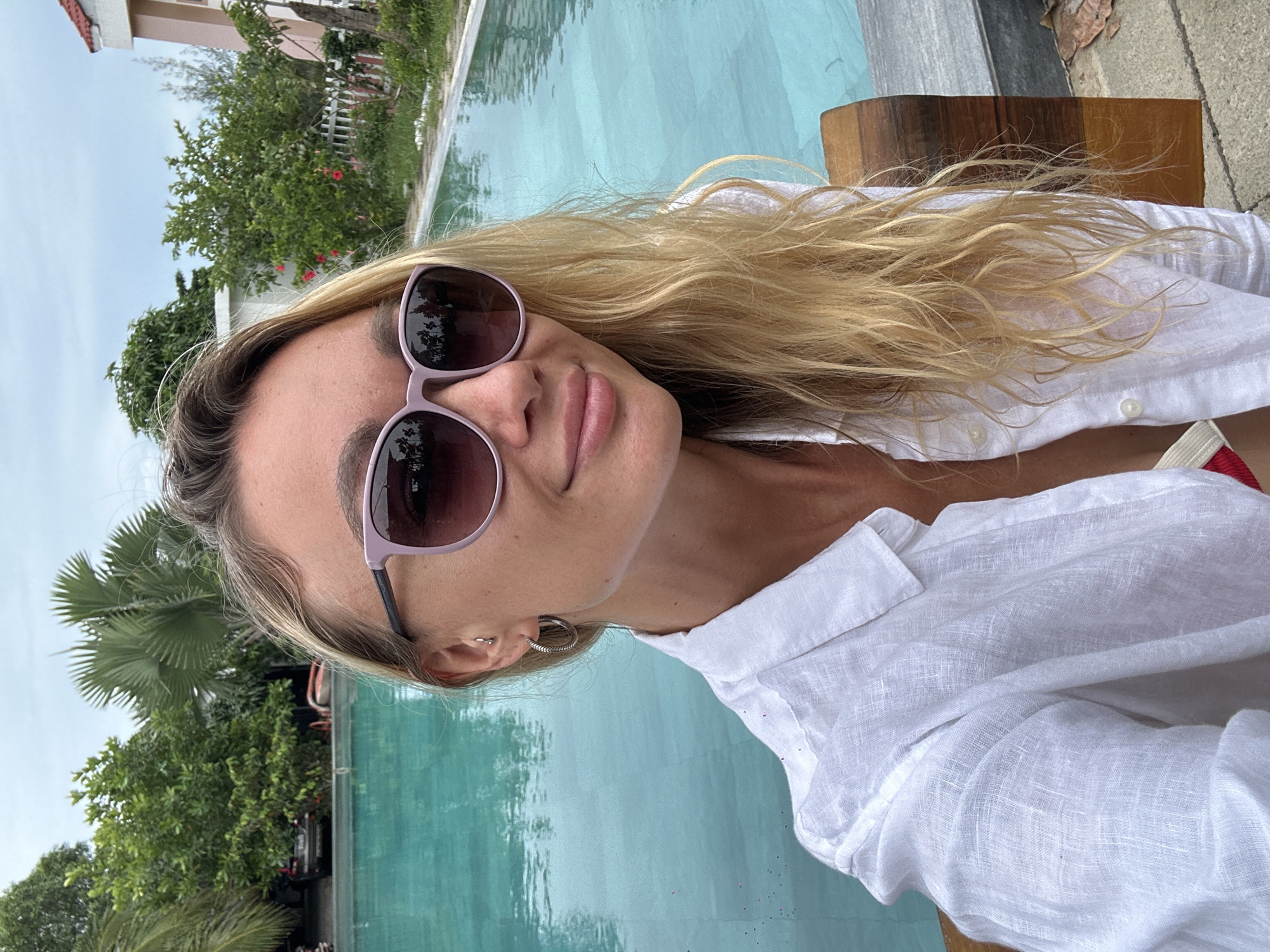 A woman with blonde hair, wearing sunglasses and a white linen shirt, smiles for a selfie by a swimming pool in Mũi Né, Vietnam. The pool, with clear turquoise water, takes up a large portion of the background. Lush green trees and palm trees surround the pool, and a pink building is visible in the distance. A wooden bench is positioned to the right of the woman.