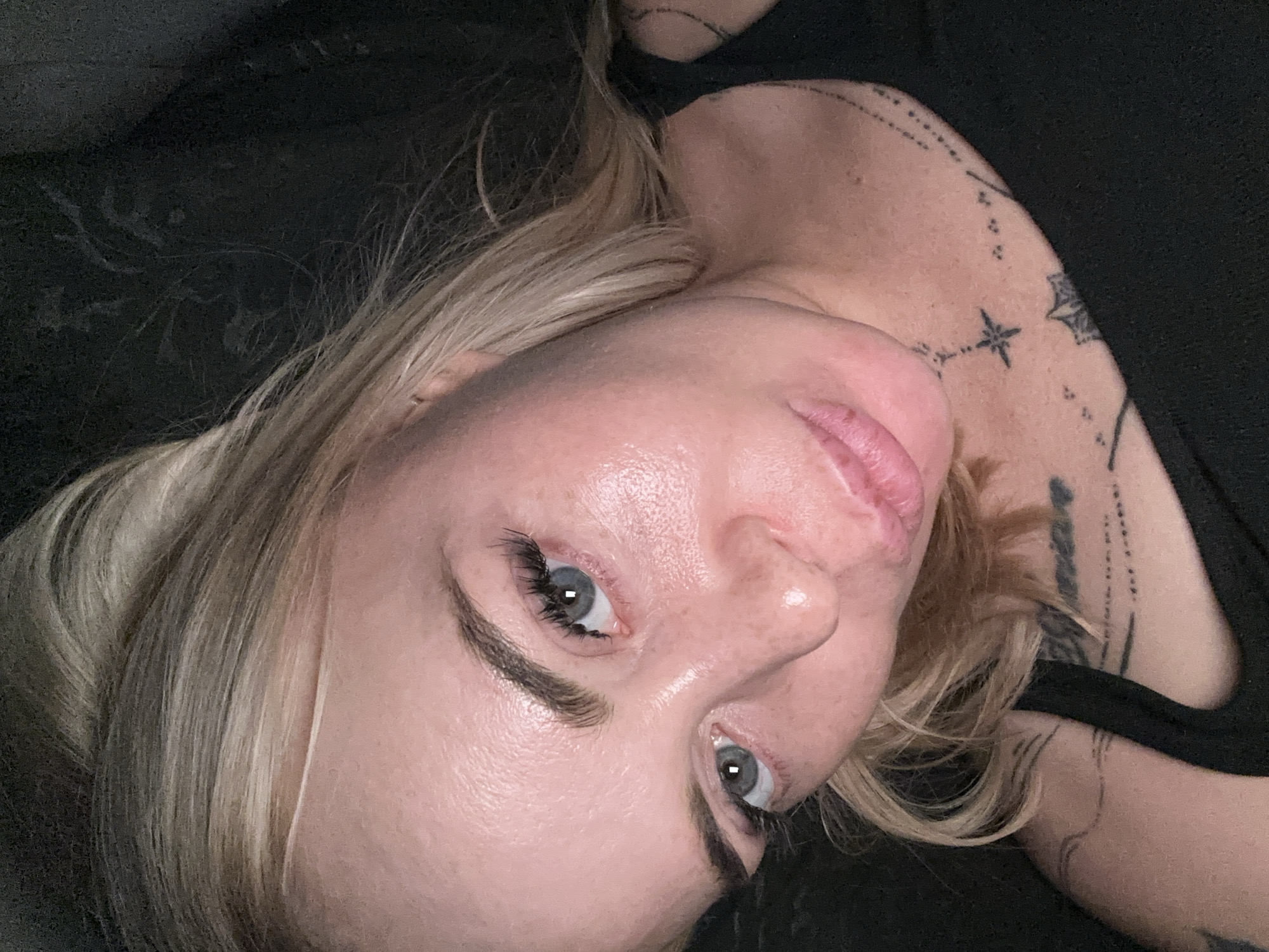 A close-up, overhead shot shows a fair-skinned person with blonde hair and blue eyes. They are wearing a black tank top with several tattoos visible on their chest and shoulder. The person's face is angled downwards and to the left, with their lips slightly parted. The background is dark and out of focus, with the lighting highlighting the person's face and chest.