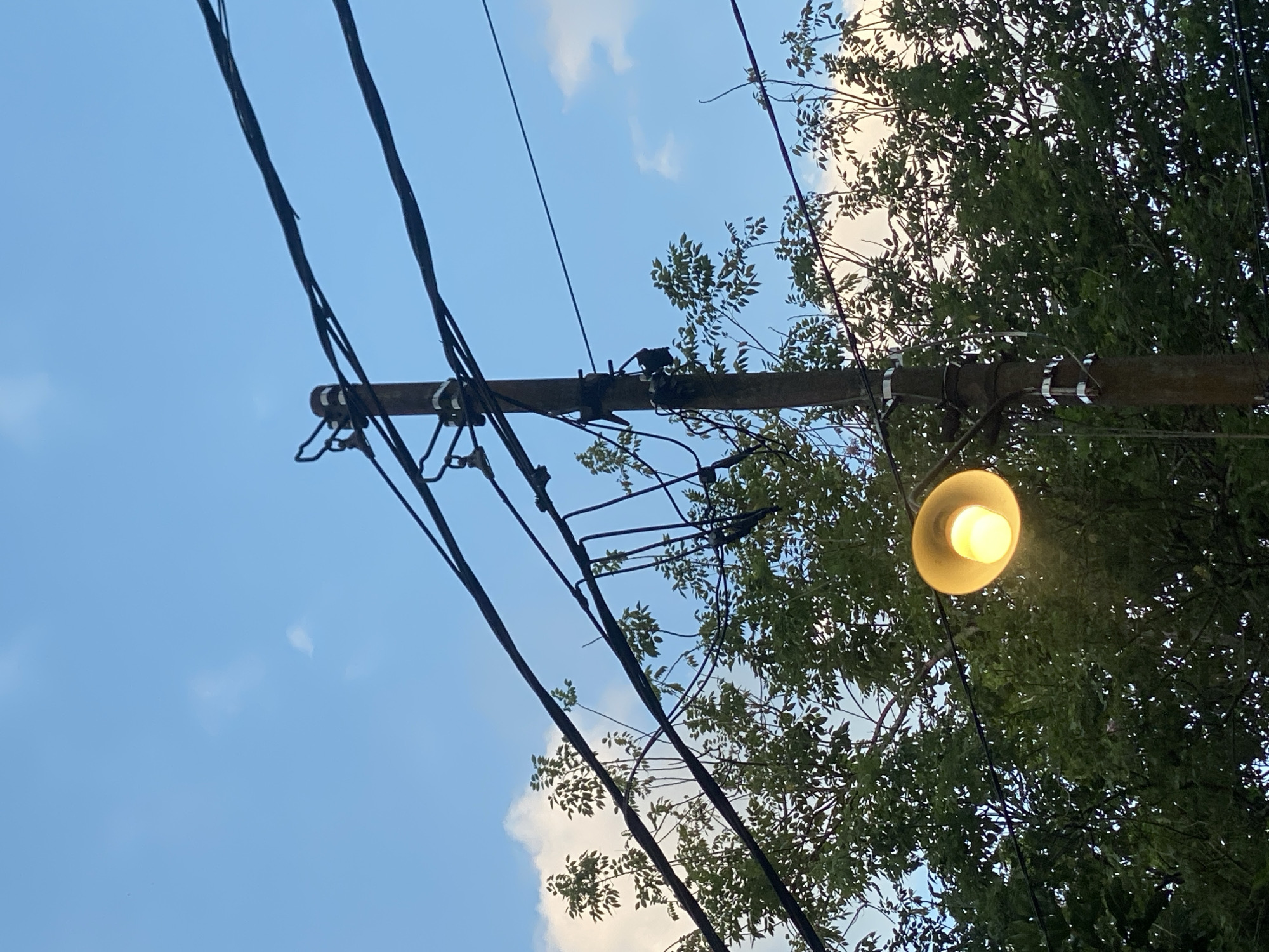 This image depicts a utility pole with power lines and a streetlamp against a blue sky with scattered clouds. The pole is made of wood and has multiple wires attached to it. A single streetlamp is illuminated with a warm, yellow light, casting a glow on the surrounding trees. The trees are lush and green, suggesting a natural or semi-urban environment. The scene appears to be outdoors, likely in a residential area or along a street in Banjarbaru, Indonesia, as indicated by the presence of such infrastructure and vegetation. There are no people visible in the image.