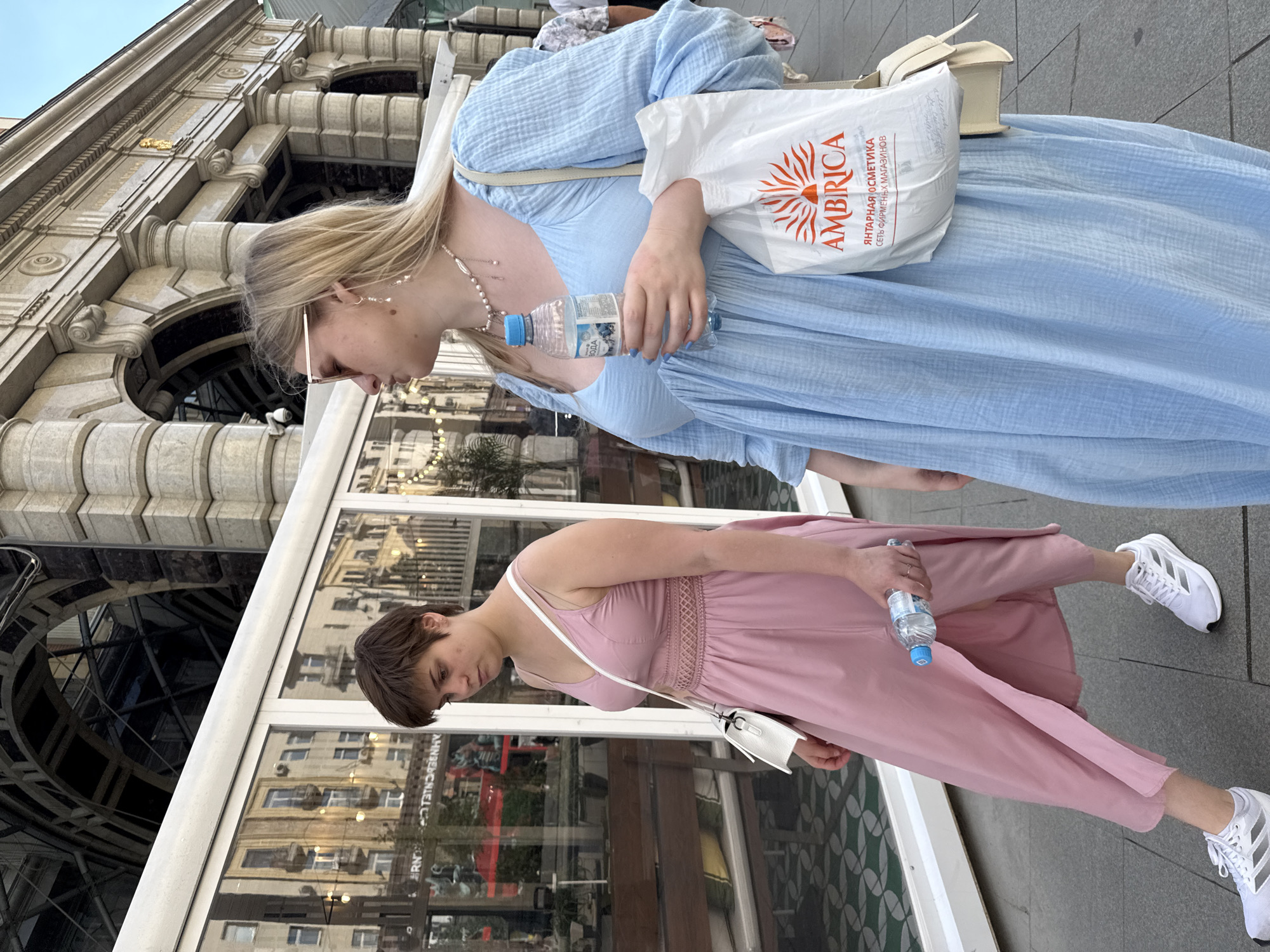 Two young women walk side-by-side on a paved city sidewalk. The woman on the left, with short brown hair, wears a sleeveless pink dress with a V-neck and a white crossbody bag. She holds a bottle of water. The woman on the right, with long blonde hair, wears a light blue, long-sleeved dress and sunglasses. She carries a white shopping bag with an orange and red sunburst logo that reads "AMBRICA" in Russian, and a beige shoulder bag. Both women are wearing athletic shoes. Behind them, the scene shows reflections in large glass windows of buildings with ornate stone facades and arched architectural features, characteristic of Moscow's historic city center. The overall impression is that of a casual outing in a bustling urban environment.