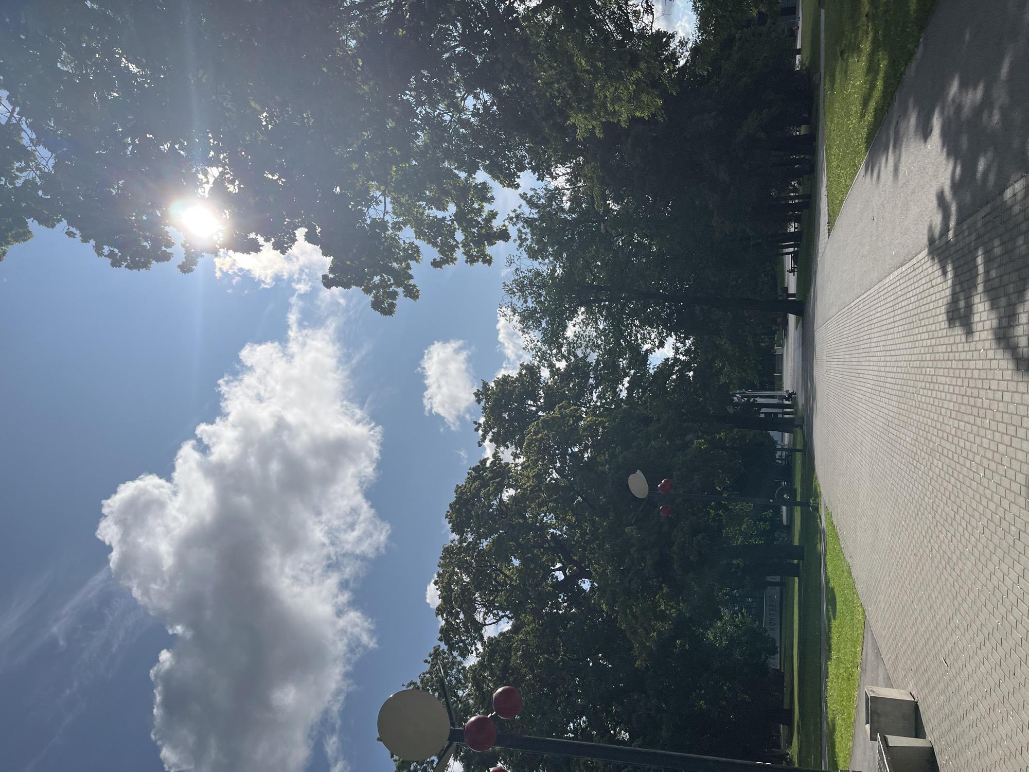 The image depicts a sun-drenched park walkway in Jõhvi, Estonia, on a clear day with scattered white clouds. A paved walkway, made of grey rectangular bricks, leads into the distance. On the left side of the walkway, there are two concrete benches. Beyond the walkway, to the left, is a grassy area with scattered trees. To the right of the walkway, also a grassy area is visible with trees and a pathway. Several tall trees with dense green foliage line both sides of the walkway, casting shadows on the path. The sun is shining brightly, with rays filtering through the leaves of a large tree in the upper right portion of the image. Two streetlights with spherical white lamps and red accents are visible on the left side of the walkway. In the distance, beyond the trees, there appears to be a building with the Cyrillic word "КОНТЕНТ" visible on it. A person can be seen sitting on a bench in the distance, to the right of the walkway.