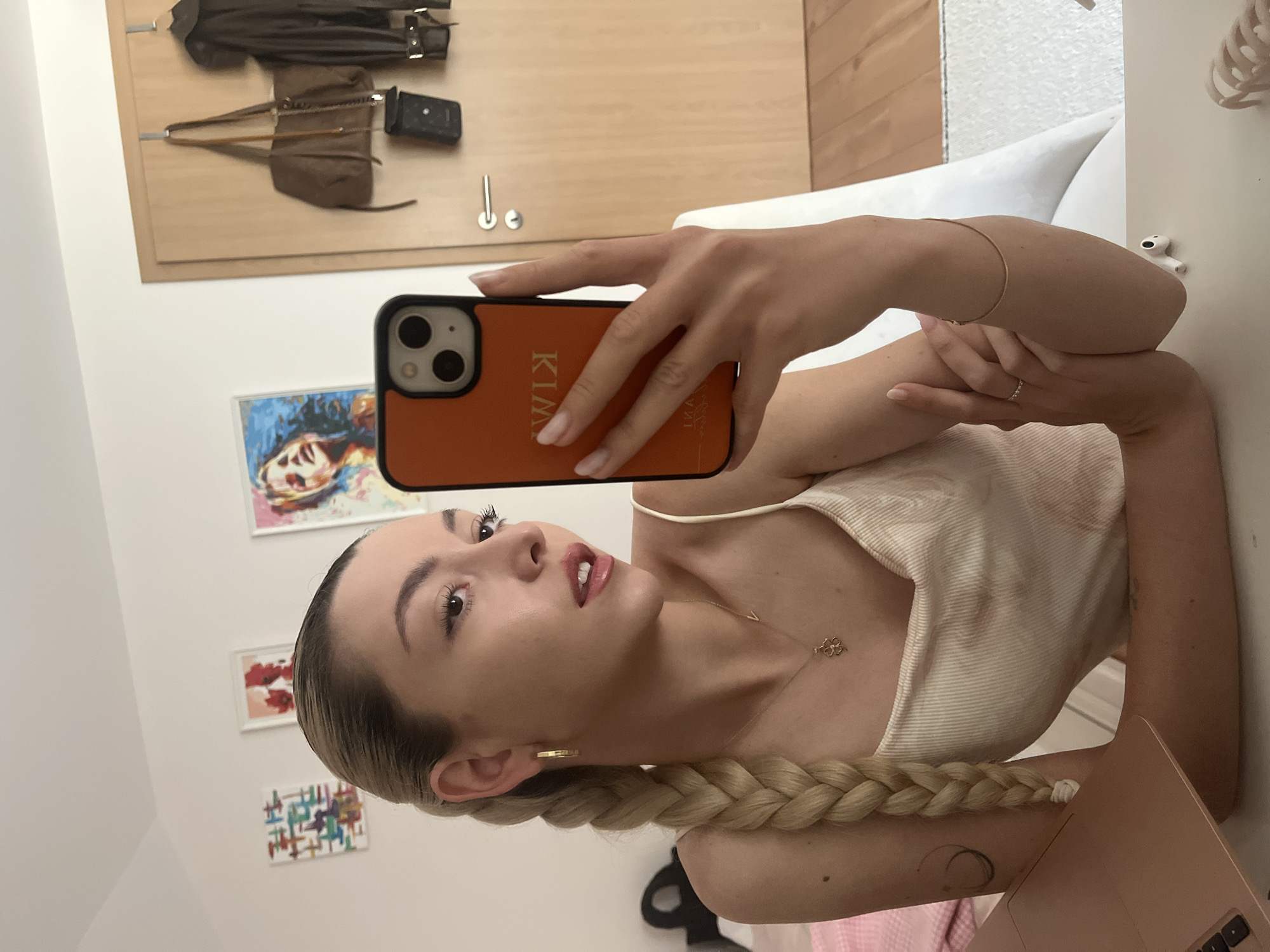 A blonde woman is taking a selfie with an orange phone case that has the name "KIM" on it. She is wearing a white tank top and gold jewelry, including earrings, a necklace, and a bracelet. Her hair is in a long braid. In the background, there is a door with a jacket, bag, and small purse hanging on it. There are also three framed pictures on the wall. The scene appears to be inside a home. The city is Unknown, Unknown.