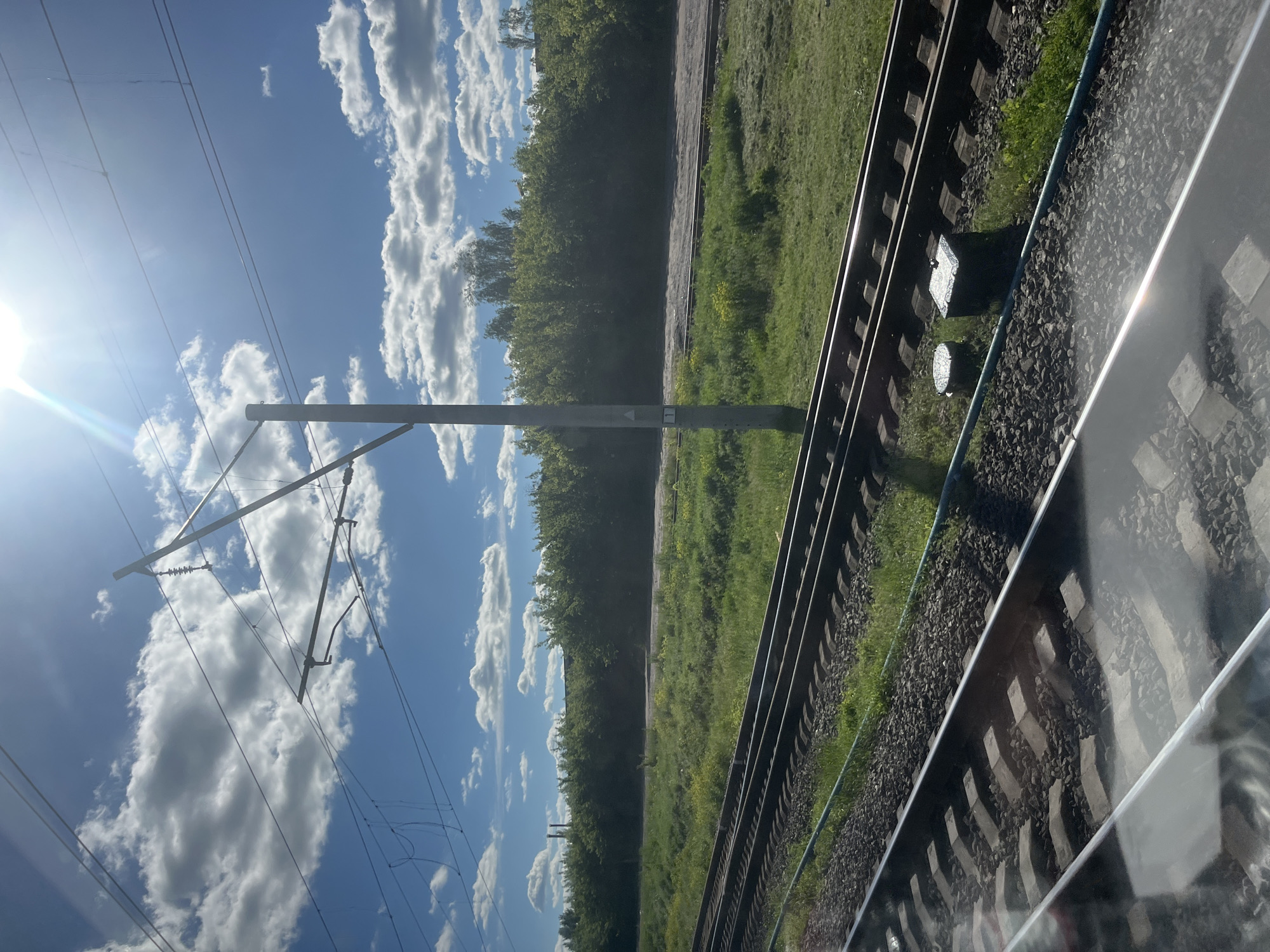 A low-angle, medium shot captures a view of train tracks against a backdrop of lush greenery and a bright, cloud-dotted sky. The sun, positioned high and to the left, casts a lens flare across the image. The foreground shows multiple sets of parallel train tracks, composed of dark ballast, concrete sleepers, and steel rails. A faint blue hose runs along the side of the tracks.
In the middle ground, a tall, grey utility pole stands beside the tracks, supporting overhead electrical wires for a train. The ground adjacent to the tracks is covered in green grass, interspersed with small yellow flowers. Beyond the grassy area, a dense line of trees forms a dark green hedge. In the distance, hints of industrial structures with chimneys can be seen peeking above the trees, suggesting the outskirts of Trylisy, Ukraine, possibly near an industrial zone or the edge of the settlement. The sky is a vibrant blue, filled with fluffy white clouds, and the overall lighting indicates a sunny daytime setting.