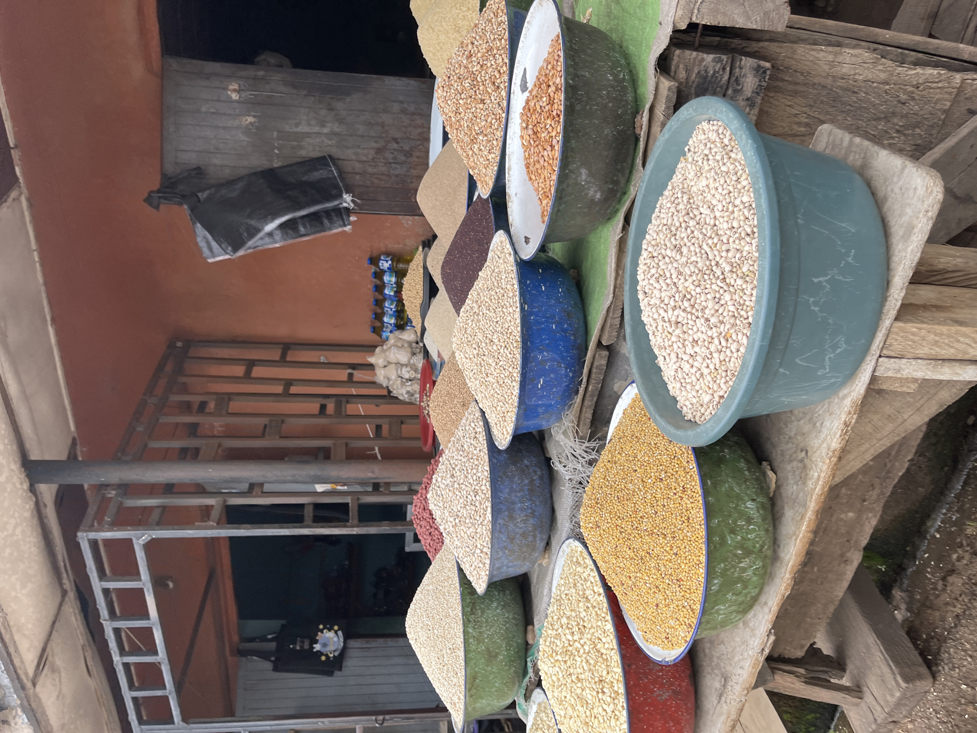 A market stall in Barakin Akawo, Nigeria, displays an array of dried grains and beans in large, colorful basins. The stall is set up on wooden planks, with a corrugated metal roof overhead. In the foreground, several basins are filled with different types of food items. A large green basin holds a mound of light-colored beans. To its left, a red-rimmed basin is filled with small yellow grains, likely millet or corn. Another large green basin, further to the right and slightly behind, contains a heap of pale, oval-shaped beans. In the middle ground, blue basins hold more grains and beans, including a large blue basin filled with light brown grains and another with pale white beans. Behind these, a red basin is filled with peanuts. Further back, additional basins and piles of various grains are visible, including a dark reddish-brown pile, likely sorghum or a similar grain, and light brown grains. The backdrop consists of a building with orange-colored walls and a metal structure. A black plastic bag hangs from the wall. Bottles of liquid and more piles of grains are visible inside the stall. The ground appears to be dirt or gravel.