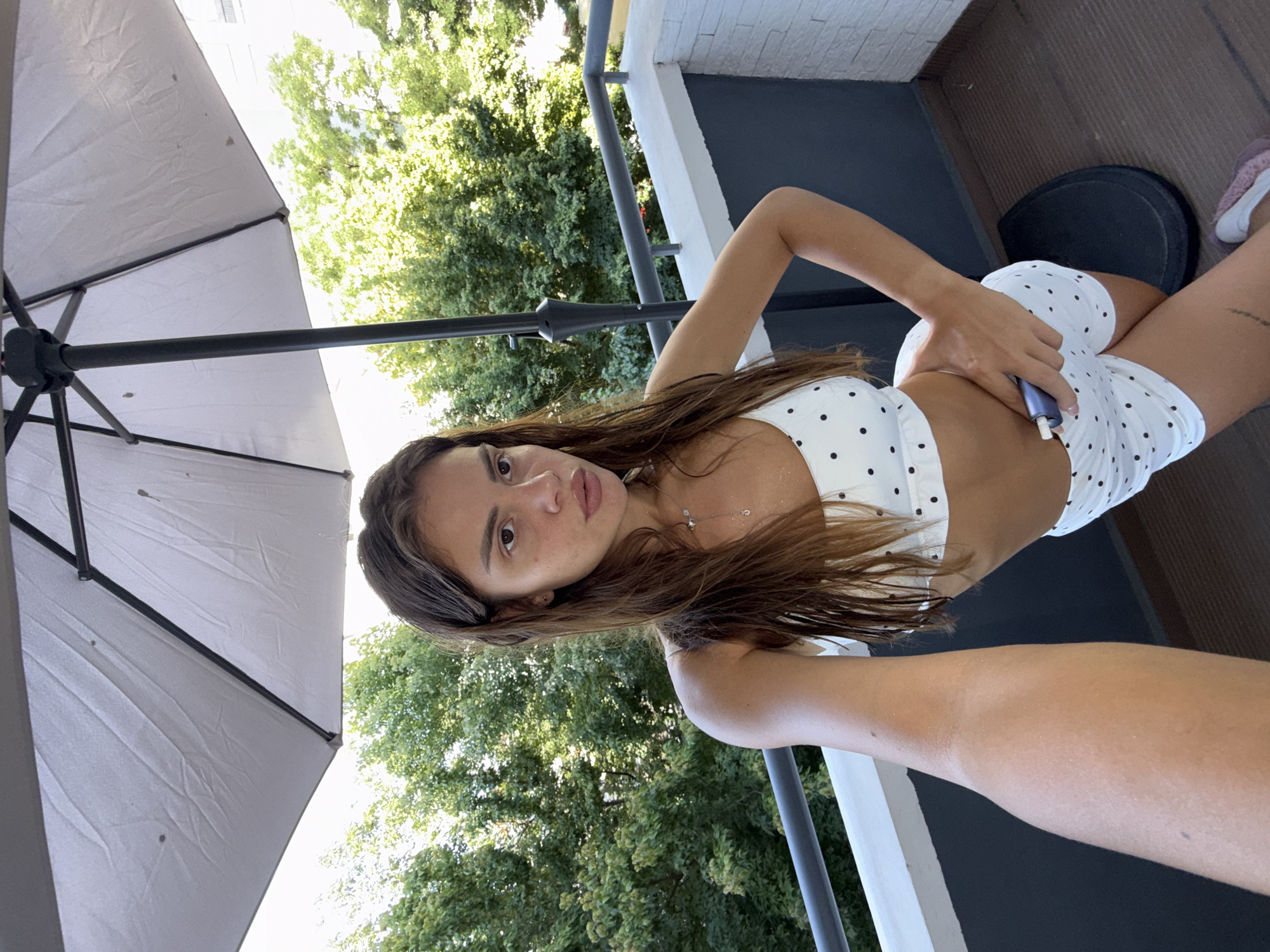 A young woman with long, brown hair is taking a selfie on a balcony in Plovdiv, Bulgaria. She is wearing a white bikini-style top and shorts with black polka dots. She has her left hand on her hip and her right hand holding a small, cylindrical device, possibly an e-cigarette or vape pen. Behind her, lush green trees are visible, suggesting a sunny day. A large, grey umbrella stands behind her, casting some shade. The balcony railing is a combination of grey metal and white concrete. The floor of the balcony is a dark, textured material. The overall scene depicts a relaxed, outdoor moment.