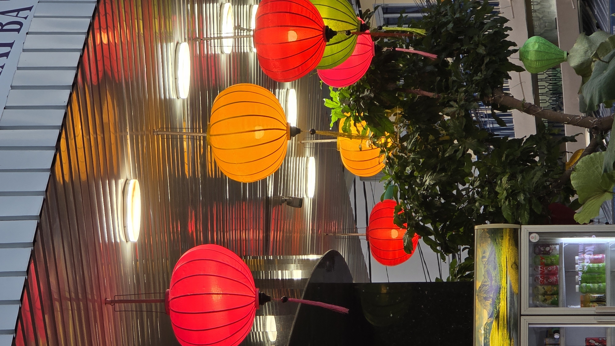 The image shows a street scene in Nha Trang, Vietnam, at night. Lanterns in red, orange, green, and pink hang from trees and wires overhead. A building with vertical, illuminated stripes is visible on the left. A refrigerator filled with drinks is on the right. Dark foliage is intertwined with the lanterns. The image is taken from a low angle, looking up at the lanterns and the surrounding scene.