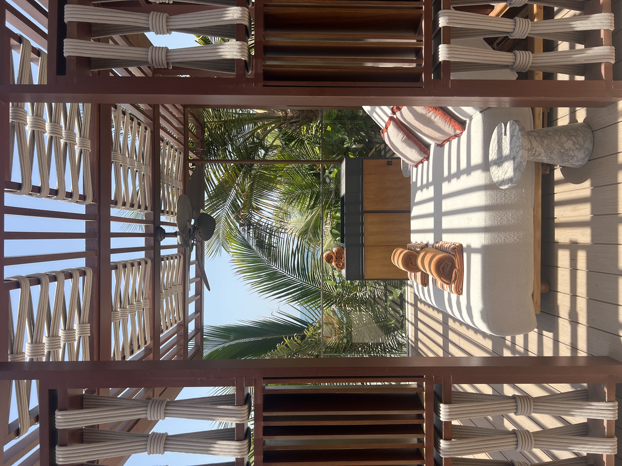 The image shows a luxury outdoor lounge area framed by a wooden structure with rope detailing. The perspective is from within the structure looking out towards palm trees and a blue sky, suggesting a tropical or resort-like setting.
Within the frame, there is a daybed or lounging sofa with cushions and rolled towels, accompanied by a small marble table. A wooden cabinet or storage unit is visible near the daybed.
The location appears to be in Dubai, United Arab Emirates, suggested by the combination of palm trees, modern architecture, and luxurious setting.