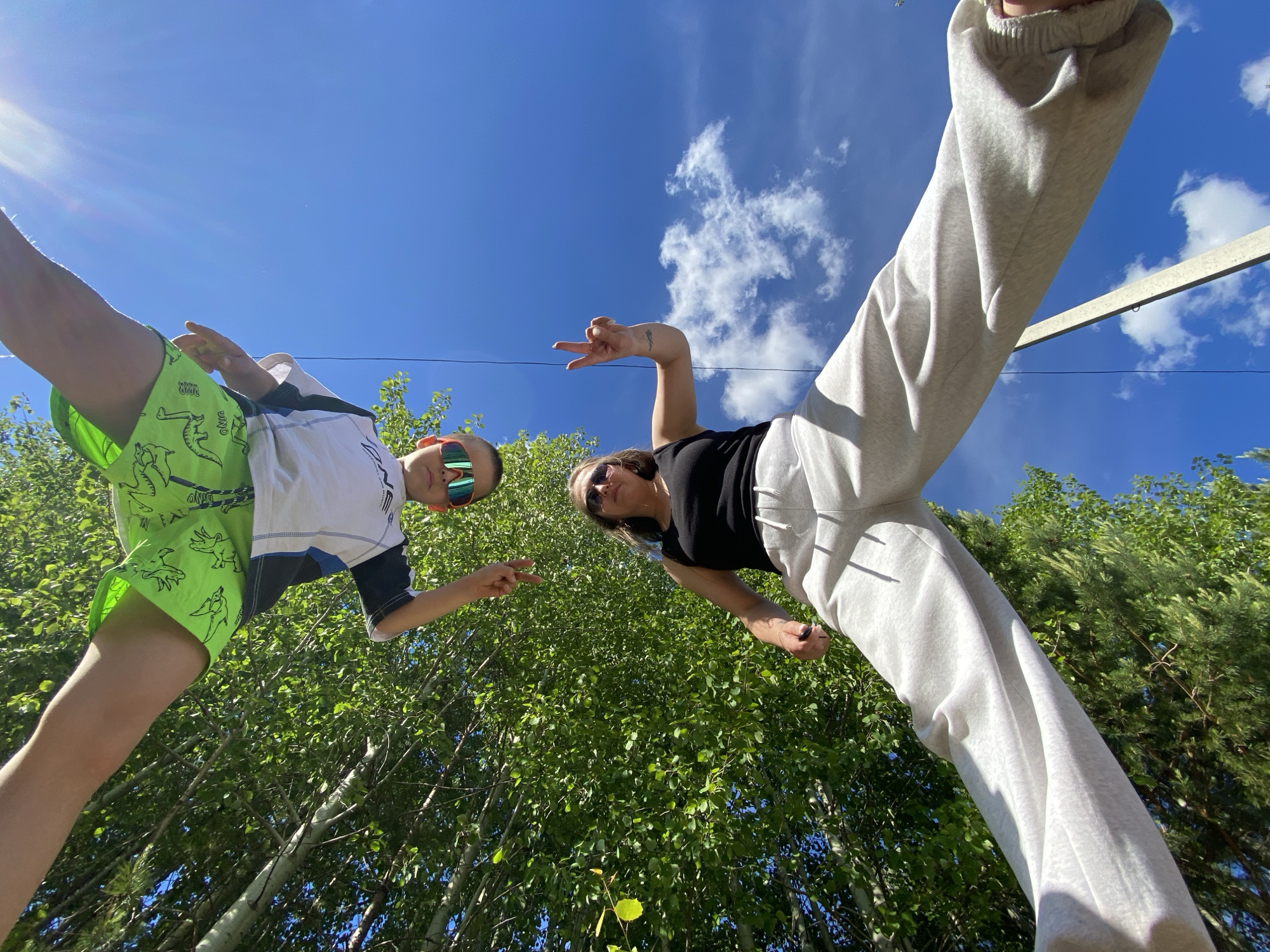 Two people are standing and looking down at the camera in an outdoor setting. On the left is a young person wearing sunglasses, a white shirt with blue and black accents, and bright green shorts with a dinosaur pattern. On the right is a person with sunglasses, a black top, and light grey sweatpants, making a peace sign with one hand. The background features a blue sky with scattered clouds above a dense area of trees. An aluminum pole runs across the top right of the image, partially obscured by trees. The photograph was taken in Chaikovsky, Russia.