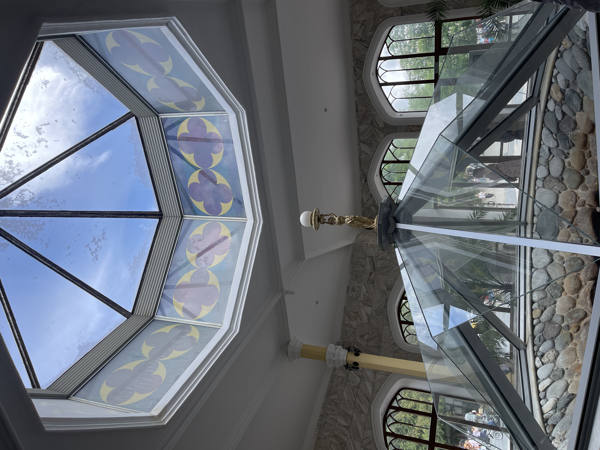 The image, taken from a low angle, presents an interior architectural view, likely a ceiling and skylight. A large, octagonal skylight with patterned stained glass panels and clear glass sections reveals a view of the sky. A central, decorative light fixture hangs beneath the ceiling. Adjacent to the skylight, are arched windows offering views of the exterior, including trees and possibly buildings.
A glass structure with metallic supports stands out, and through the glass, a garden or outdoor area with stones and greenery is visible. Reflections in the glass suggest the presence of people and objects outside. The ceiling and walls are primarily white with some sections featuring a marble-like pattern.
The city location is Kislovodsk, Russia.