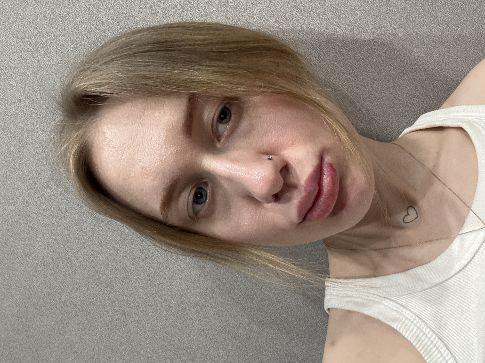 A close-up, head-and-shoulders portrait depicts a young adult female in an indoor setting in Rostov-on-Don, Russia. The subject has light blonde hair, light skin with some visible redness on the forehead and cheeks, and blue eyes looking directly at the viewer. A small, shiny stud pierces her left nostril. Her lips are full and slightly pouted, with a small red lesion visible on the lower lip near the left corner. She wears a white ribbed tank top and a thin gold-colored necklace featuring a small, black outline heart pendant situated on the sternum. The background consists of a plain, light grey wall with a subtle, textured finish.