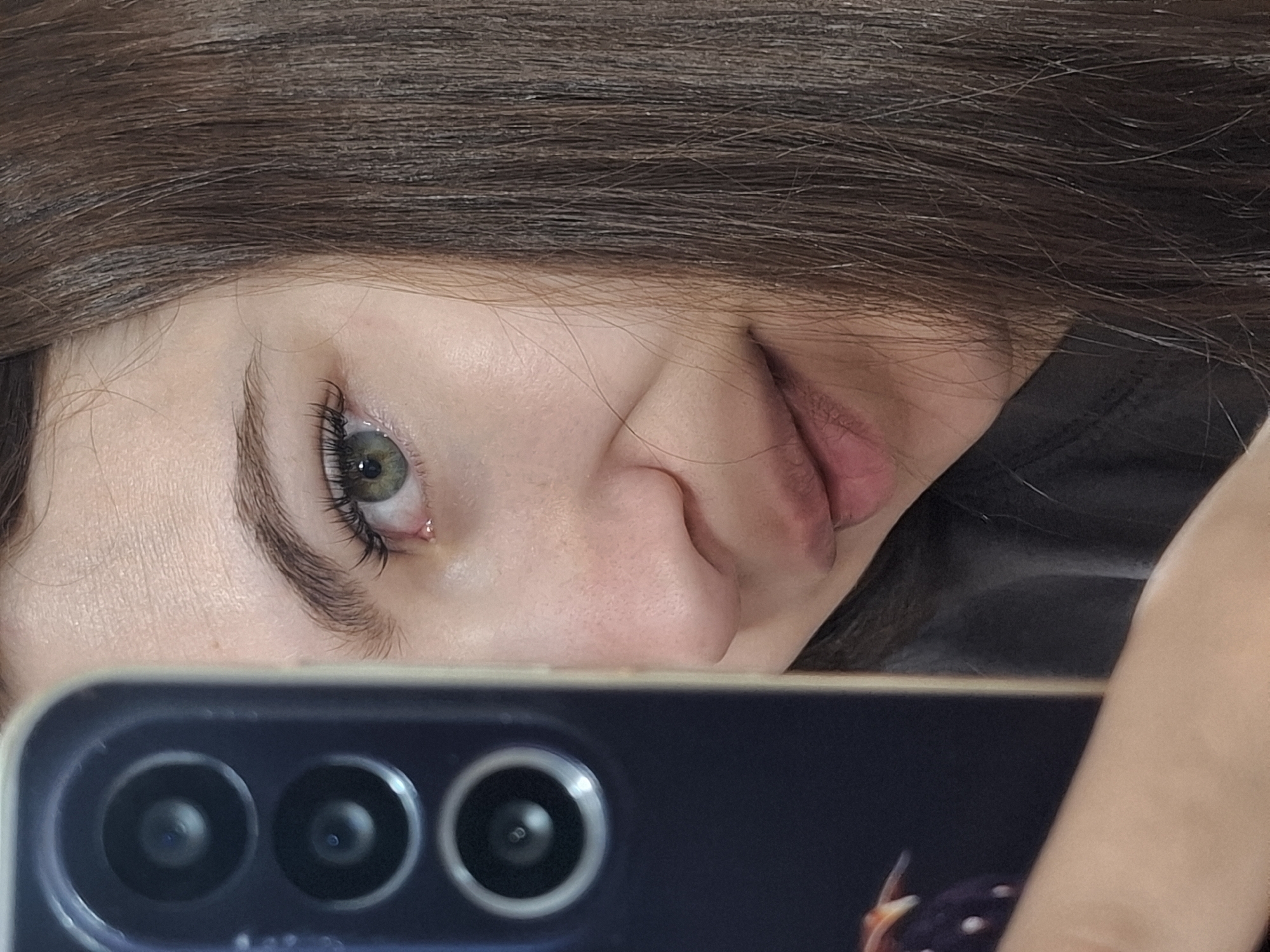 A close-up selfie shot reveals a young woman's face partially obscured by a smartphone. Her face is tilted to the left, with light brown hair falling across the top of the frame. She has fair skin, a small nose, and full lips. Her left eye is green with long eyelashes. She is looking at the camera and pouting slightly.
The smartphone is positioned in the foreground, partially covering the bottom of the frame. Three camera lenses are visible on the back of the phone, arranged horizontally. A fragment of a design or object can be seen on the phone's back.
The background is out of focus.
There is no information about the location in the city of Chaikovsky, Russia.