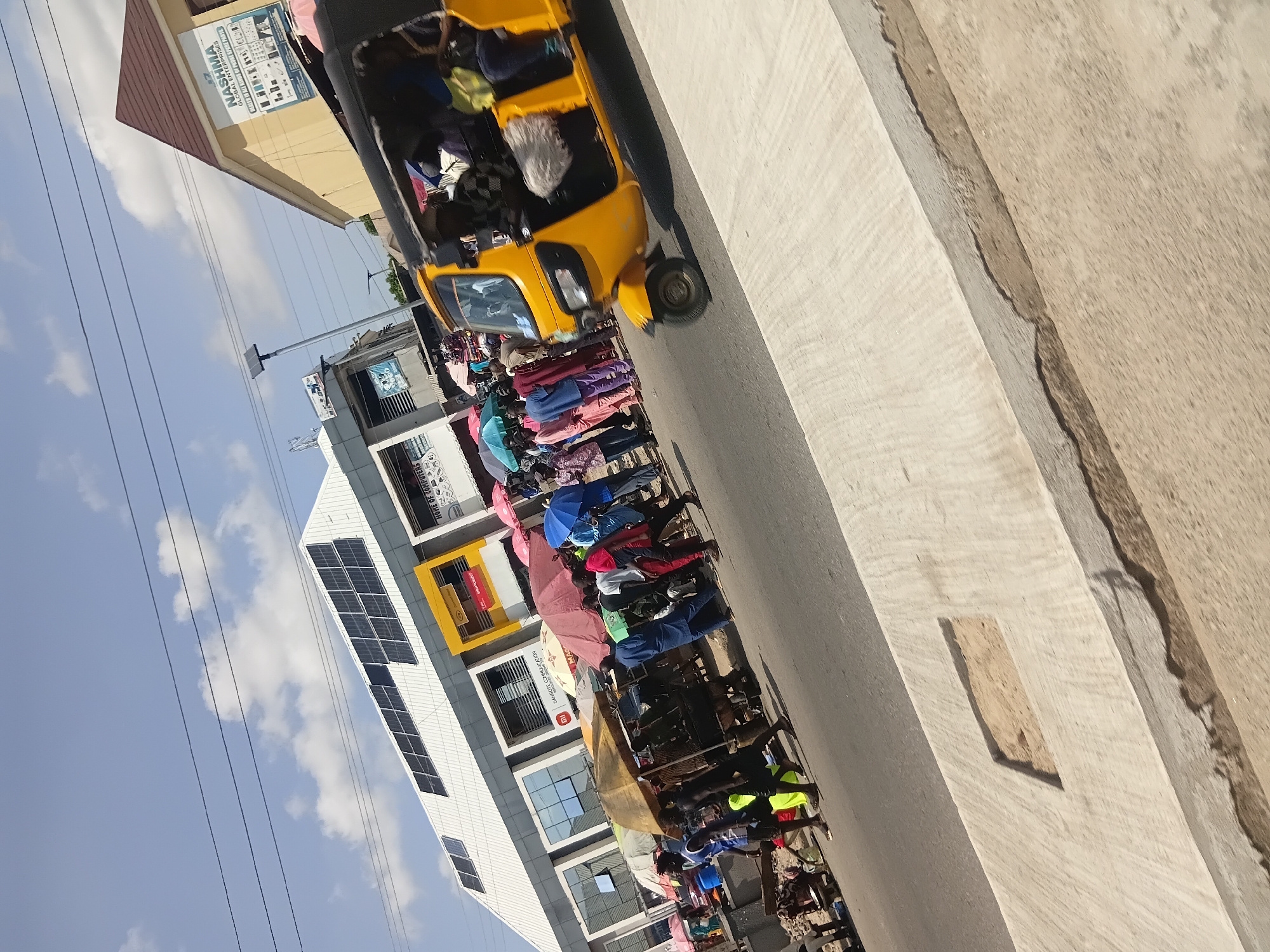 In Jalingo, Nigeria, a vibrant street scene unfolds under a bright, partly cloudy sky. The foreground features a paved walkway with a recessed rectangular section filled with sand. To the right, a yellow auto-rickshaw, a common form of transport in the region, is captured in motion, with passengers visible inside.
The middle ground depicts a bustling market area with numerous people gathered under colorful umbrellas, seeking shade from the sun. Stalls are set up, suggesting trade and commerce. Various individuals are seen walking and interacting, some carrying goods.
In the background, a modern building with solar panels on its roof stands out, contrasted by an older, yellowish building with signage indicating "NASHMA GLOBAL ENTERPRISES" and a list of services. The overall impression is one of lively urban activity in Jalingo.