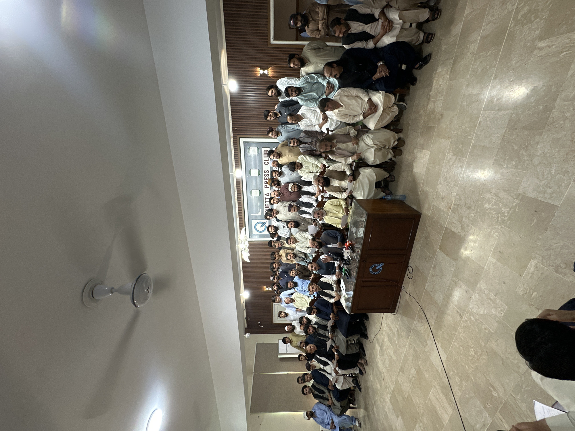 A large group of men, likely over 50, are seated in an indoor space at the Quetta Press Club in Quetta City Tehsil, Pakistan. The men are mostly wearing traditional clothing, including shalwar kameez and some turbans. They are arranged in rows facing a wooden podium from which microphones are visible, suggesting a press conference or public statement is about to be made or has just concluded. A sign with "QUETTA PRESS CLUB" can be seen on the wall behind the group. The room has tiled flooring and a ceiling fan.