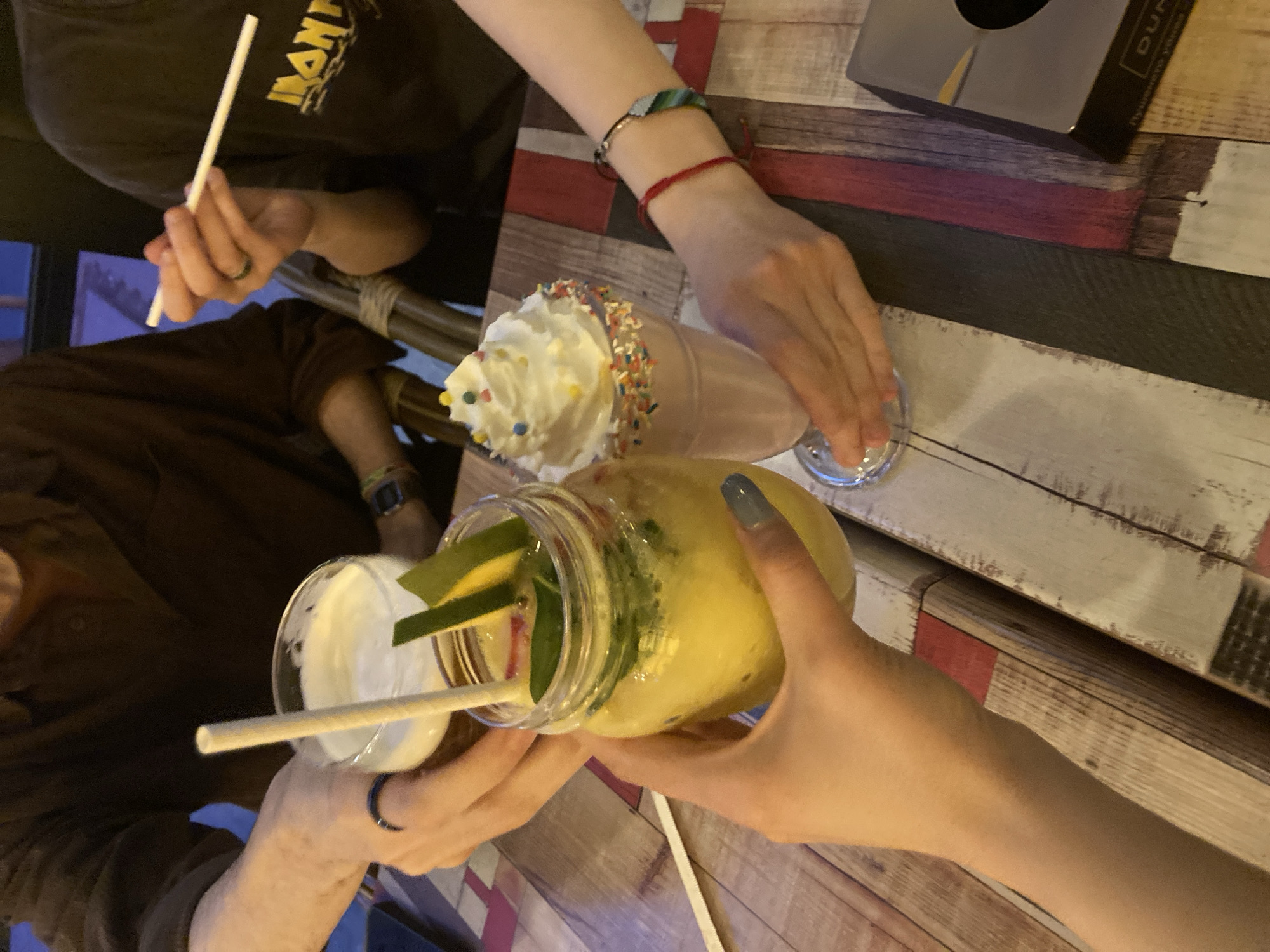 The image shows a wooden table setting with three individuals. Two individuals are holding drinks - one with a yellow liquid in a mason jar with fruit slices and a straw, the other with a white creamy drink in a glass. There is also a milkshake with whipped cream and sprinkles on the table. All individuals are seated. A straw is also visible near the hand holding the mason jar. A black box is on the upper right corner of the image. No information about the city or the location is available from the image.