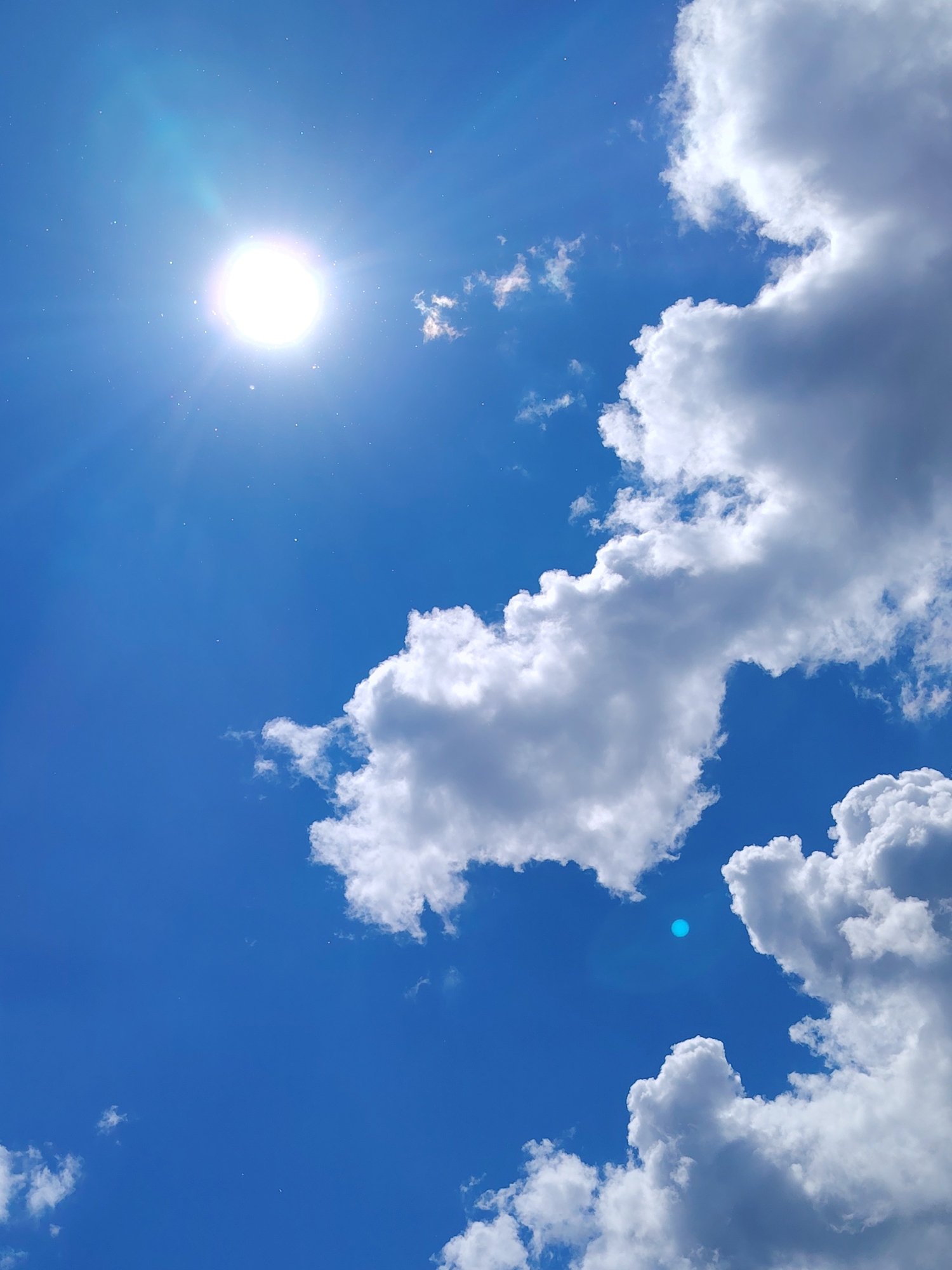 The image shows a daytime sky, likely in Chaikovsky, Russia, with a bright sun in the upper left quadrant, casting rays. Fluffy white cumulus clouds are present in the right half of the frame, contrasting with the clear, deep blue sky. A lens flare is visible beneath the clouds. There is no indication of people, specific objects, or actions. The environment is a clear sky with scattered clouds.
