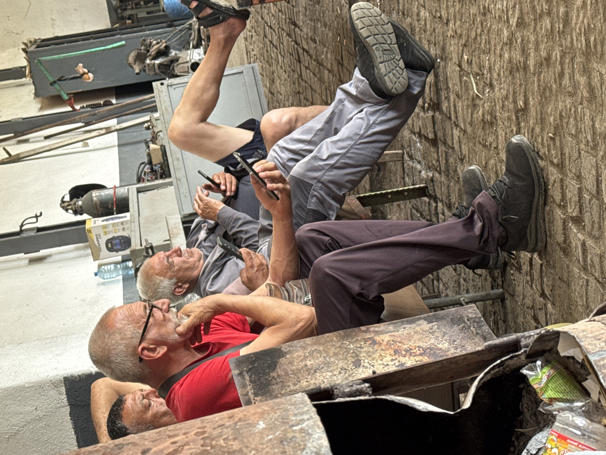 The image captures four men relaxing in what appears to be an informal workshop or storage area. The scene is somewhat industrial and rustic, with a worn, textured floor and rough walls.
In the foreground, a man with a grey beard and glasses, wearing a red t-shirt, reclines with his arm resting on a rusty metal structure, looking upwards. Next to him, partially visible, is another man with dark hair, also appearing to be at rest.
Further back, two other men are seated, both with grey hair, deeply absorbed in their smartphones. One wears a grey long-sleeved shirt, holding a black phone, with his legs clad in light grey trousers and black shoes extended forward. The other man’s legs, in dark pants and black sneakers, are propped up against a textured, brick-like wall or structure. The legs of a fourth man, wearing blue shorts and black sandals, are visible in the upper background.
The setting includes various industrial elements: metal frames, machinery, and a gas cylinder are discernible in the background. A plastic water bottle sits on a shelf. In the immediate foreground, rusty metal containers, one holding discarded packaging including a green snack packet and a red-and-white label fragment ("PAP S."), contribute to the unkempt, working environment aesthetic. The men appear relaxed and disengaged from their surroundings, focused on their phones, suggesting a break or a period of leisure during the day.