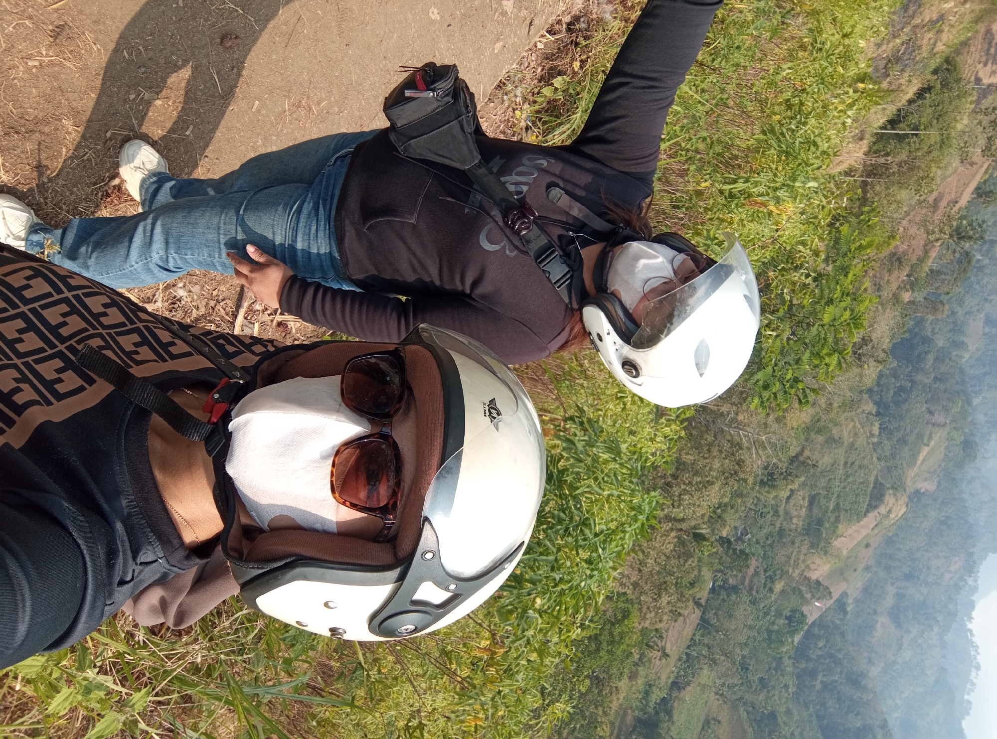 In Pandansari Lor, Indonesia, two individuals wearing white helmets and white face masks pose for a photo on a dirt path overlooking a lush, terraced hillside. The person in the foreground, positioned slightly to the right, wears sunglasses and a black shirt with a patterned strap crossing their chest. The second individual, to the left and further back, has their left arm extended, possibly taking the photo. This person wears a black hoodie over a white shirt, blue jeans, and white sneakers, with a black crossbody bag slung over their shoulder. The background reveals a vast, green landscape with agricultural terraces, indicating a rural or semi-rural setting. The lighting suggests it is daytime, with shadows cast on the dirt path.