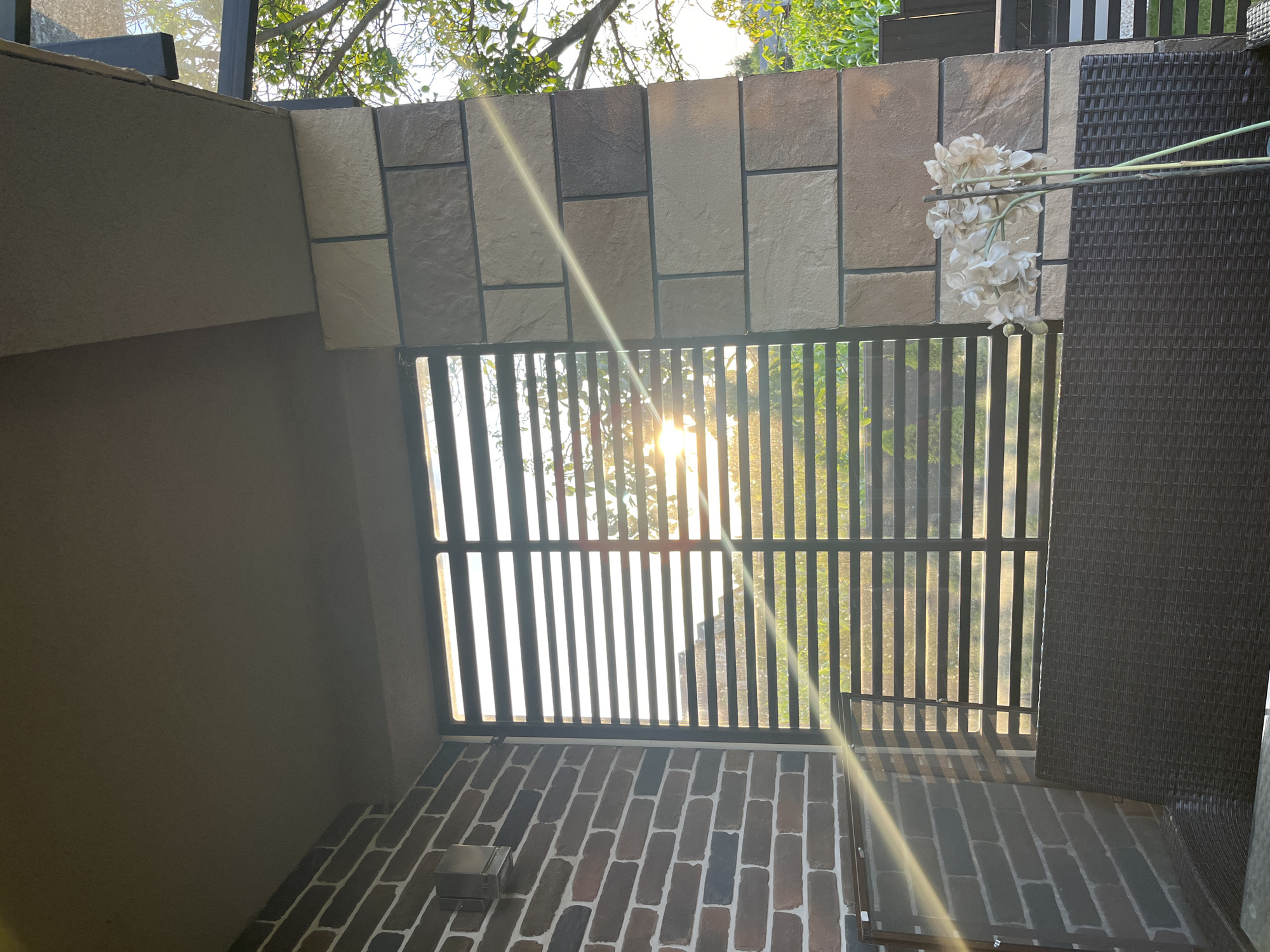 This media file depicts an outdoor setting, possibly a balcony or patio, with a view of trees and sunlight filtering through a black metal railing. The railing divides the space, with a brick floor visible below and a wall with stone tiles above. Artificial white flowers are attached to the railing on the right side. The sun is shining brightly, creating lens flares and casting long shadows, suggesting it is either morning or late afternoon. The overall impression is one of a tranquil outdoor space bathed in natural light, possibly in a residential area given the architectural elements. There is no visible text. The location is identified as Koroviia, Ukraine.