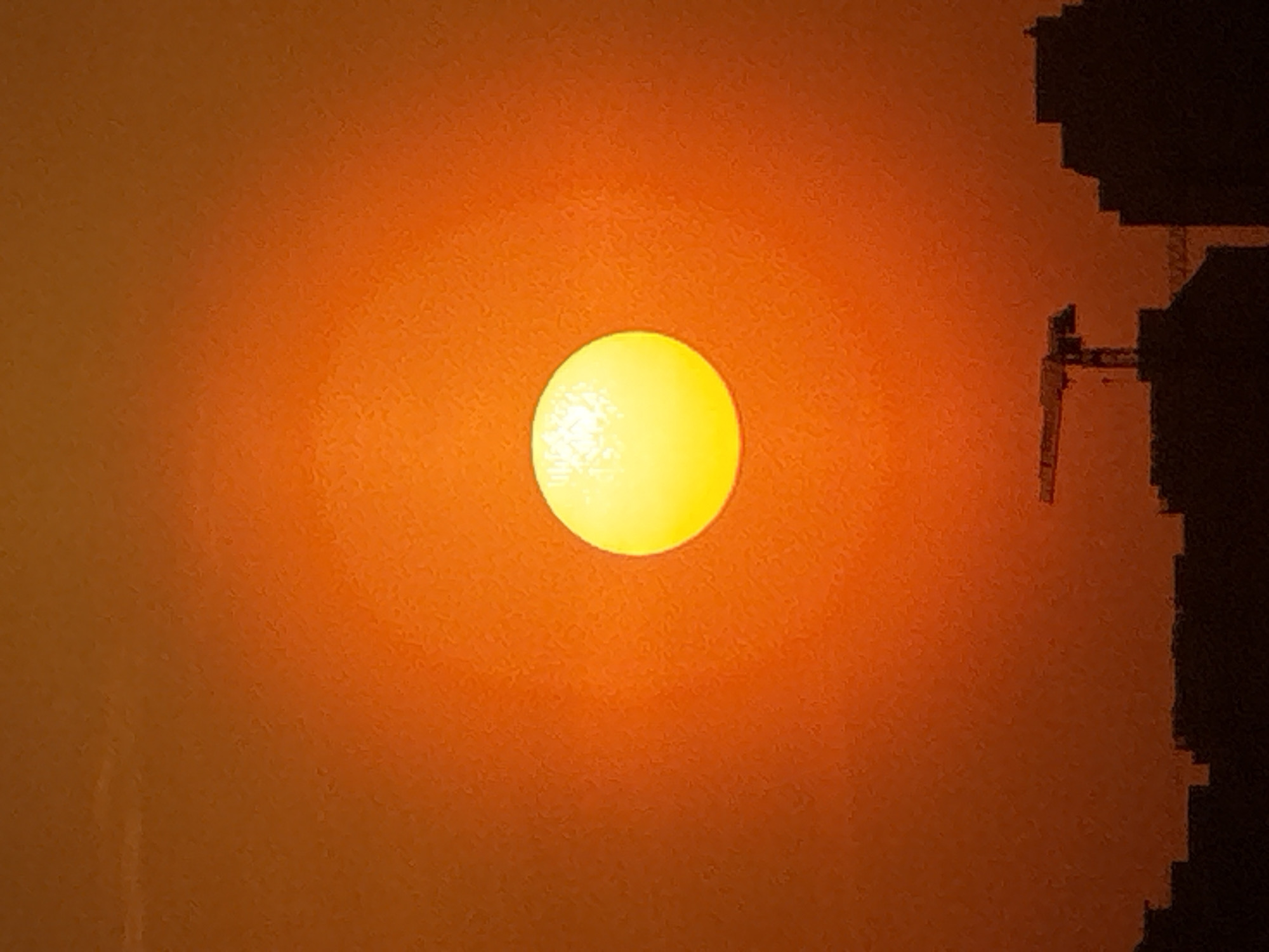 The image shows a bright yellow sun blurred by a heavy orange haze or smoke. The sun is centrally positioned. To the right, silhouetted against the orange sky, is a dark, jagged structure that resembles a building or complex. Without additional information, it is impossible to determine the specific location in Moscow, Russia. The overall atmosphere suggests a sunset or sunrise obscured by pollution, smoke, or a sandstorm.