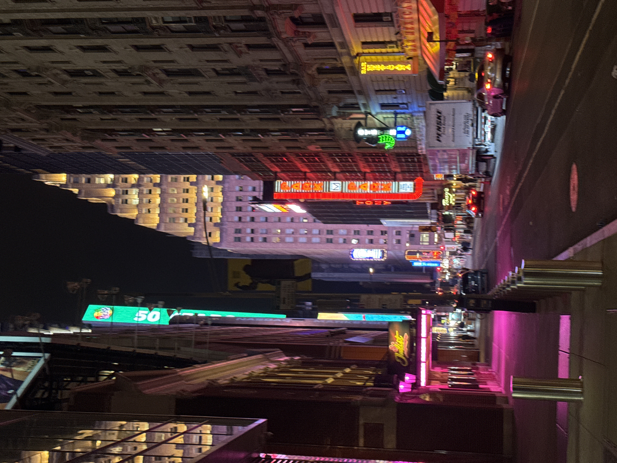 The image depicts a nighttime scene in Times Square, New York City. The view is oriented vertically. Various buildings are visible, some with lit windows and facades. Neon signs advertise businesses like "Park Lane" and "Bar Mexicano". Several vehicles are on the street, including a large truck and various cars with illuminated tail lights. Pedestrians appear to be walking on the sidewalk. There are structures with cylindrical elements, potentially part of a building's design or street furniture. The lighting creates a vibrant, colorful urban environment.