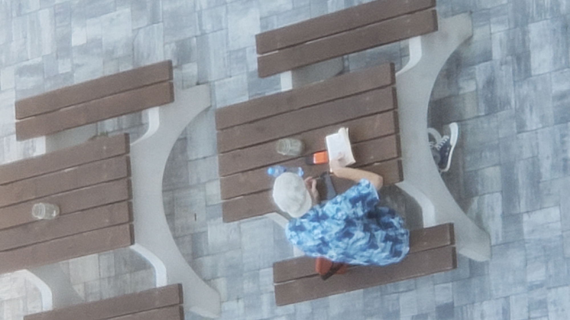 The image depicts an outdoor scene with two wooden picnic tables, each with a concrete base, set on a paved ground. The first table, in the background, has a glass jar on it. The second table, in the foreground, is occupied by a person wearing a blue and white patterned shirt and a light-colored cap. The person is seated and appears to be reading a book, with a bottle of water, a cell phone, and a jar next to them on the table. Their legs are visible beneath the table, and they are wearing dark sneakers with white soles. The environment suggests a public area, possibly a park or an outdoor seating area. The image quality is slightly grainy, and the lighting is overcast, indicating a daytime setting under diffused light conditions. The specific location in Prešov, Slovakia is not discernible from the visual information present in the image.