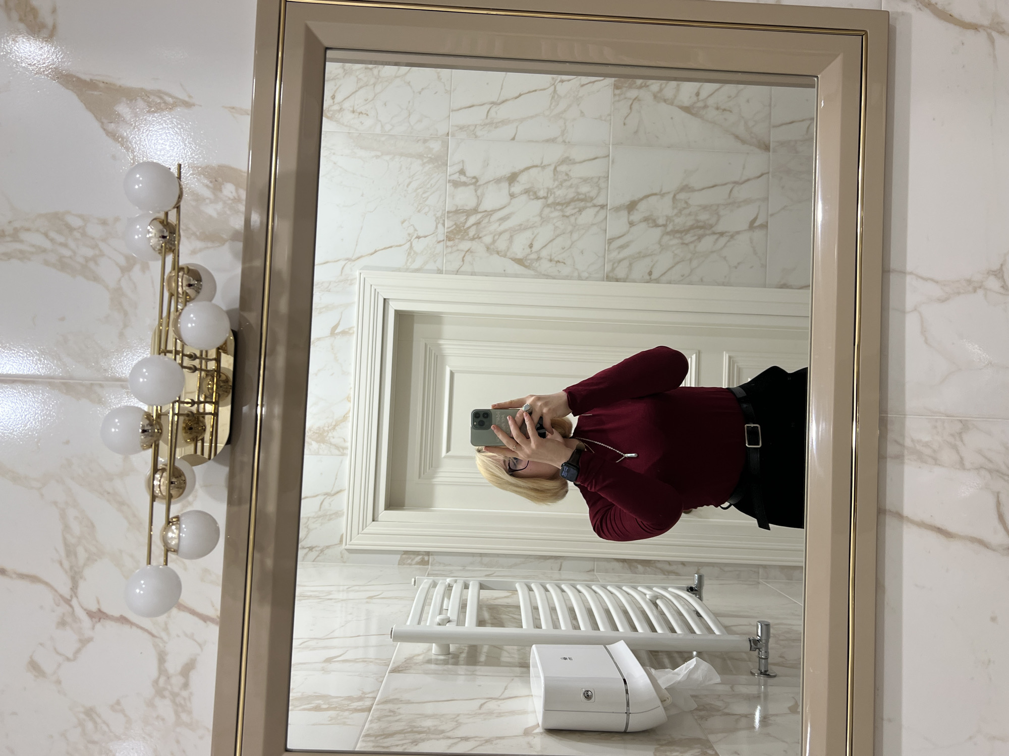 The image shows a mirror selfie taken in a modern bathroom, presented rotated 90 degrees counter-clockwise. The scene is set in Odesa, Ukraine.
Reflected in the large mirror is a person with blonde hair, wearing a dark red long-sleeved top and a black belt, capturing their reflection with a smartphone. The phone, encased in silver/grey, obscures their face. A dark, square-faced watch is visible on their left wrist, and a ring adorns their left hand.
The mirror itself features a wide beige frame accented with a thin gold line. The reflection reveals white marble-patterned wall tiles with light brown veining, which extend throughout the bathroom. A white, ornate door frame is visible in the background of the reflection. Also reflected are a white wall-mounted towel warmer or radiator and a white tissue dispenser, which has the text "LG" printed on its side.
To the left of the mirror, mounted on the matching marble-tiled wall, is a decorative gold-colored wall light fixture. It features five spherical white bulbs arranged vertically. The overall impression is of a clean, well-lit, and stylish bathroom setting.