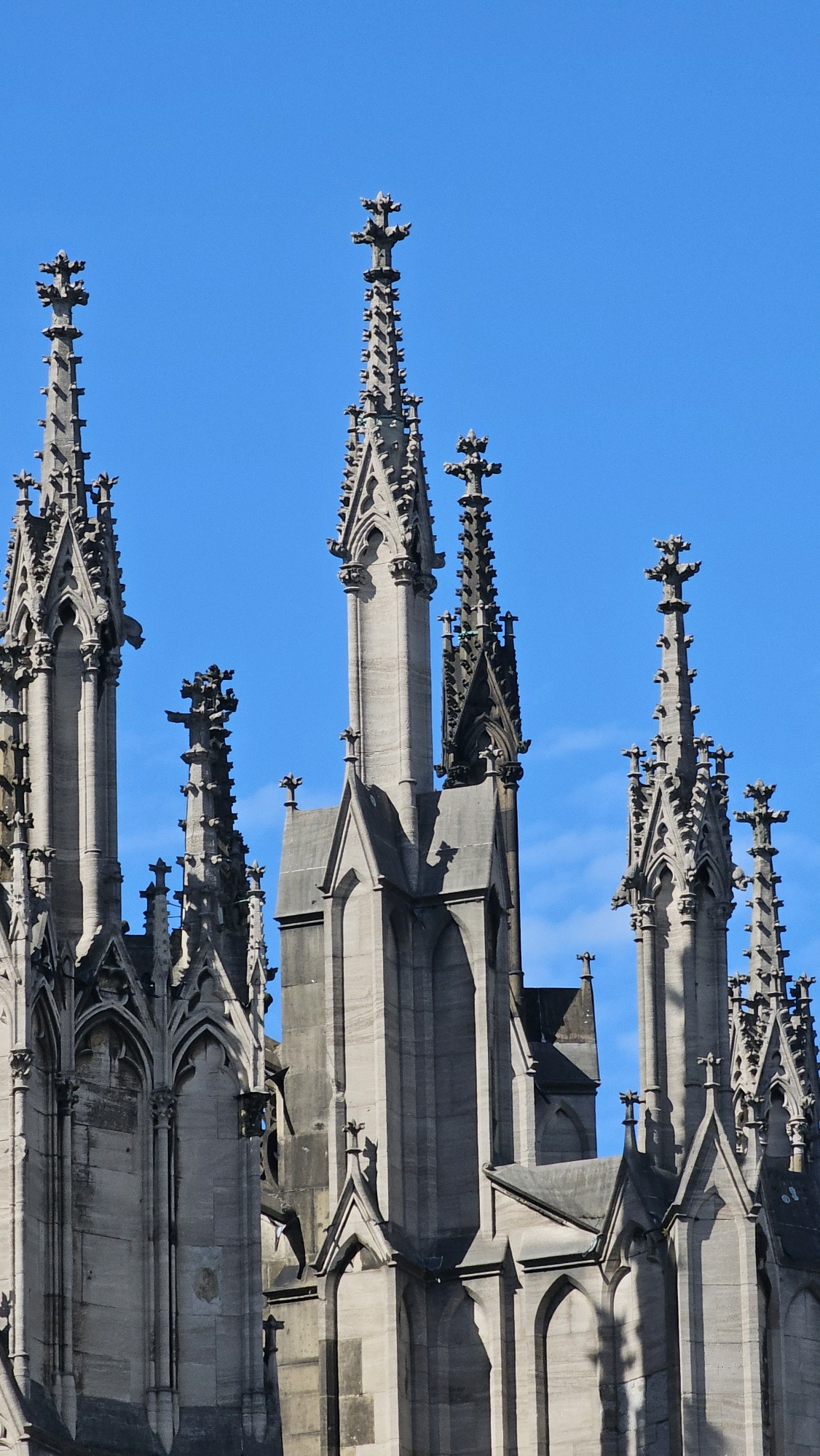 The image shows the spires of Cologne Cathedral, a UNESCO World Heritage Site located in Cologne, Germany. The Gothic cathedral's intricate stonework and tall spires dominate the frame against a clear blue sky. The architectural details, including pointed arches, gargoyles, and ornate finials, are visible. The perspective is from below, looking up at the imposing structure. No people or actions are depicted.