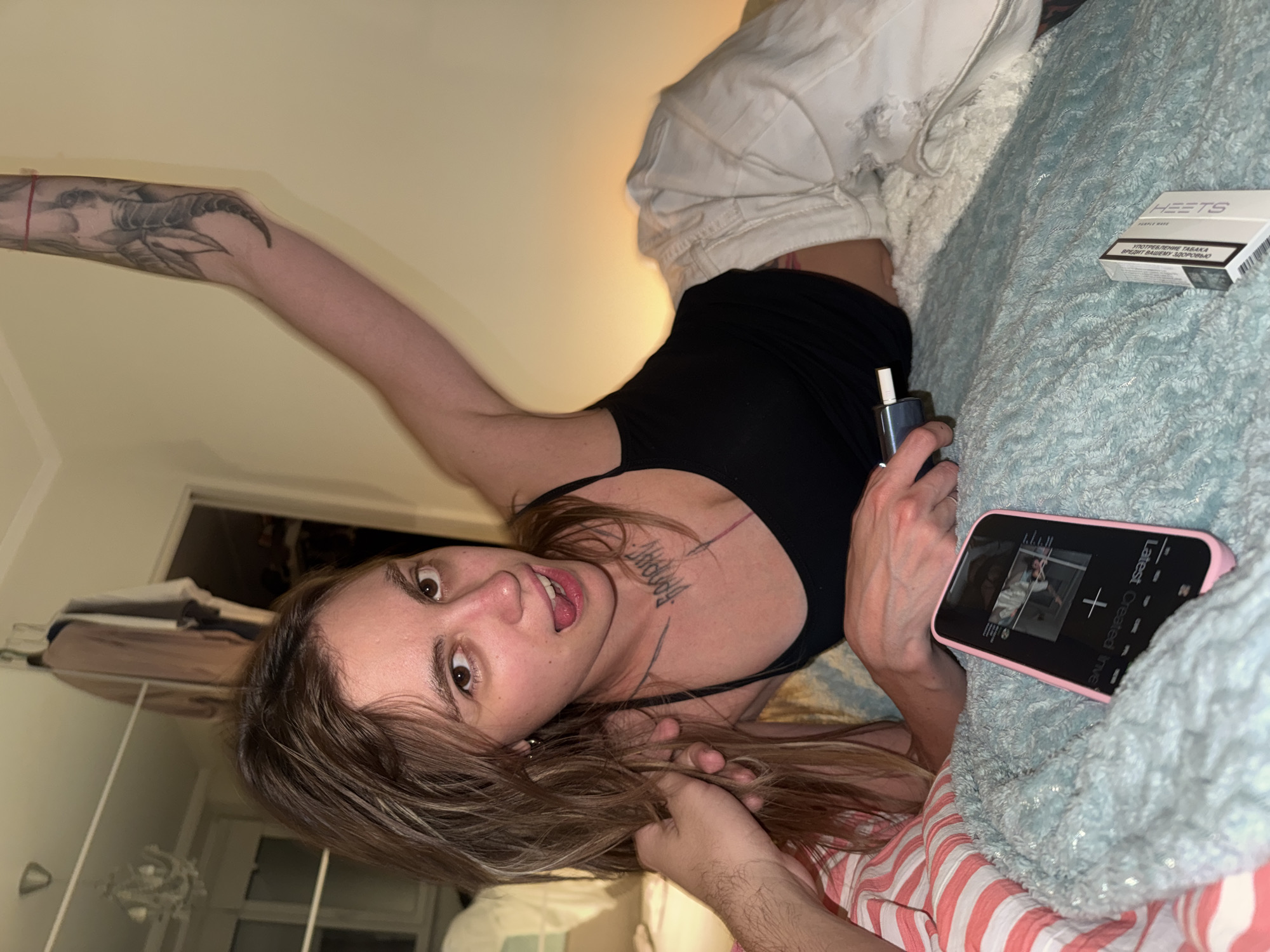 A young adult with long, light brown hair is lying on a bed with white and pink striped bedding and a light blue, textured blanket. The person is wearing a black tank top and white pants. They are looking at the camera with their tongue sticking out slightly. A hand is visible in the foreground, touching the person's hair. On the bed are a pink smartphone displaying a video, a vape pen, and a pack of HEETS cigarettes. In the background, there is a doorway leading to another room, a closet with clothes hanging, and a chandelier. The environment appears to be a bedroom.