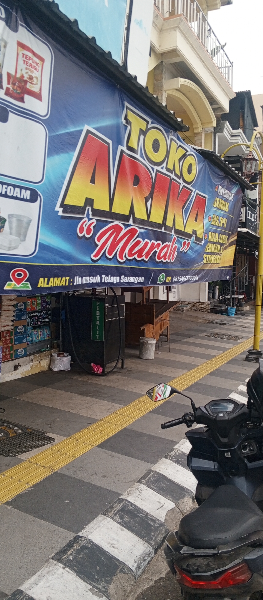 The image depicts a storefront in Sarangan, Indonesia, featuring a large blue banner with yellow and red lettering. The banner prominently displays the text "TOKO ARIKA MURAH" and includes contact information for the store, such as its address "Jln masuk Telaga Sarangan" and a phone number "HP. 081946375000". The banner also lists various products sold at the store, including "Sembako," "Gas LPG," and "Kemasan Styrofoam." To the left of the banner, a smaller graphic shows a bag of "TEPUNG TERIGU" (wheat flour) and plastic containers.
In front of the storefront, a black object with the word "PERTALIT" written vertically in green letters is visible, likely a fuel dispenser. A parked motorcycle is in the foreground, partially obscuring the sidewalk and a pedestrian tactile paving strip. The environment appears to be a street-side location, with a building facade visible in the background. The overall scene suggests a commercial area in Sarangan, Indonesia.