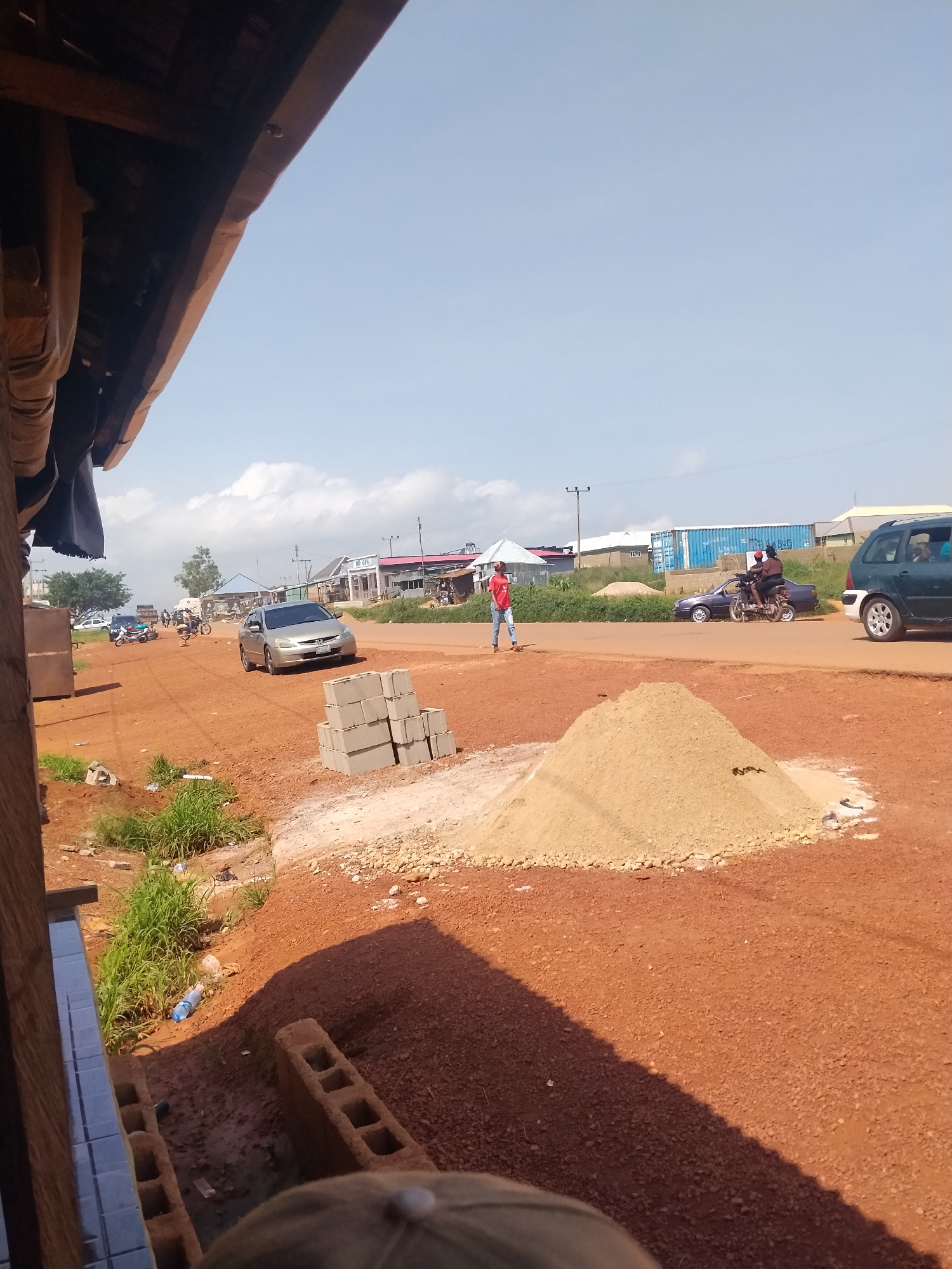 The image shows a dusty, red-dirt road in Durumi, Nigeria, with a line of low-rise buildings and a few trees in the background under a clear sky. In the foreground, a pile of sand and stacked concrete blocks indicate construction. Several vehicles are present: a silver sedan driving on the road, a dark green SUV parked on the right, a motorcycle with two riders, and another motorcycle. A person in a red shirt and blue jeans walks on the left side of the road, while another person in a black shirt and dark pants is on the motorcycle. The scene appears to be a busy street corner with ongoing construction.