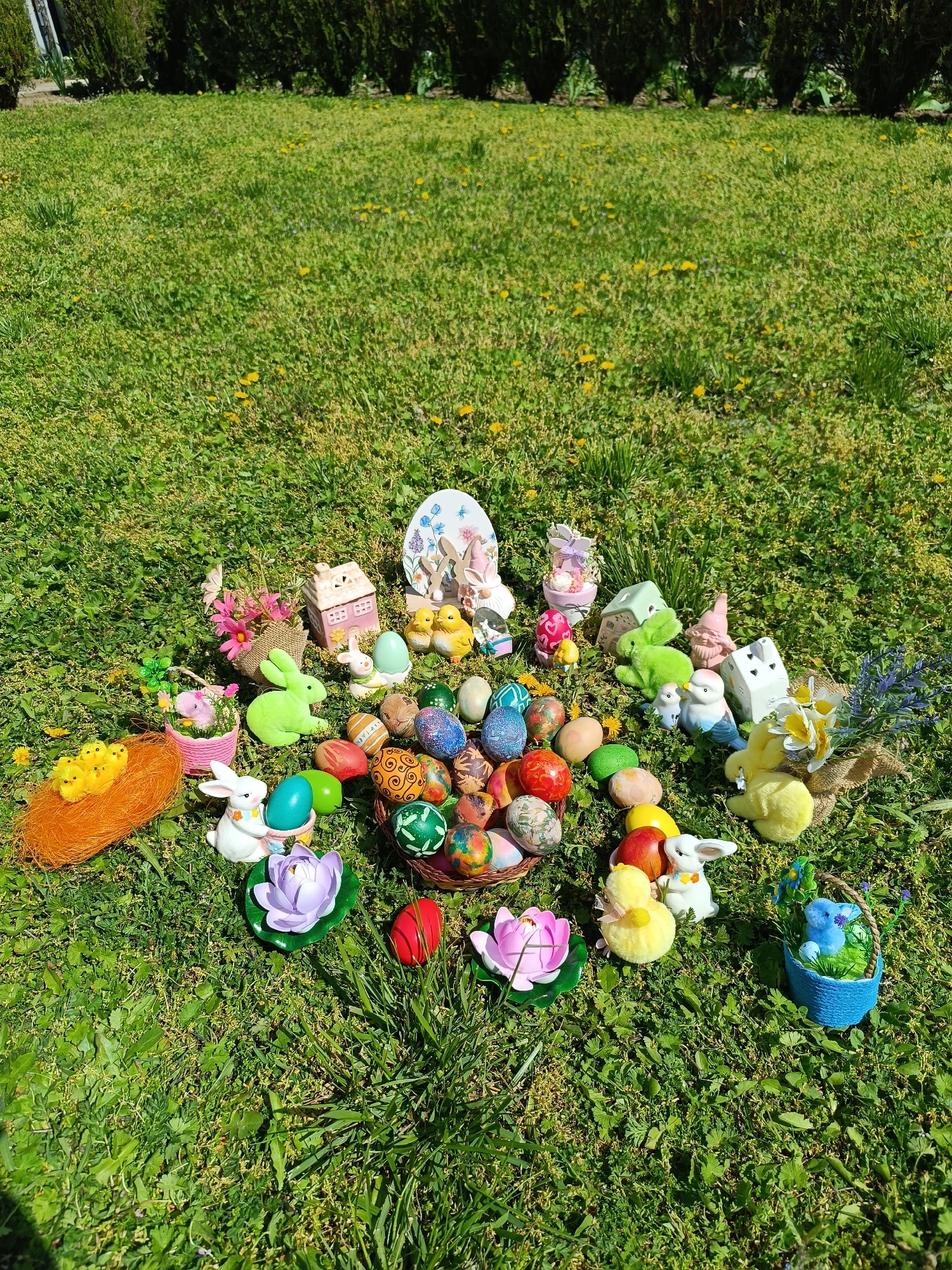 The image shows an outdoor scene on a grassy area, possibly a lawn, with various Easter-themed decorations. The decorations include a basket filled with decorated eggs, Easter bunnies, chicks, flowers, and miniature houses. The environment is likely a garden or yard, with dandelions visible in the grass. The setting is Cherkovitsa, Bulgaria. There are no people visible in the image. The overall impression is that of an Easter display or arrangement.
