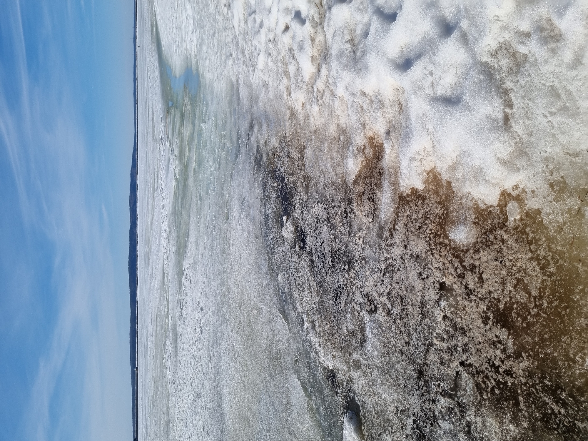 The image displays a wide-angle view of the partially thawed Votkinsk Reservoir in Chaikovsky, Russia. The immediate foreground, on the right, consists of an uneven accumulation of white snow with visible depressions. To the left, a broad, irregular area of dark brown to black shallow water, slush, and sediment-laden ice borders the snow. The midground presents an expansive, relatively flat sheet of melting ice, characterized by varied textures of bare ice, slush, and residual snow. An irregular channel of greenish-blue open water or very thin ice runs diagonally through the mid-right portion of the ice sheet, extending towards the distant horizon.
The background features a low, dark landmass, consistent with a tree-covered shore or hills, stretching across the entire horizon. On the far left of the horizon, a grey artificial embankment or breakwater extends into the water, supporting a small, white, cylindrical structure, possibly a light tower or navigational aid. A solitary, dark, conical marker object stands on the ice in the mid-distance. The sky overhead is a clear pale blue with minimal, wispy white clouds. Bright daylight illuminates the scene, consistent with late winter or early spring conditions. No individuals or explicit activities are observed within the frame.