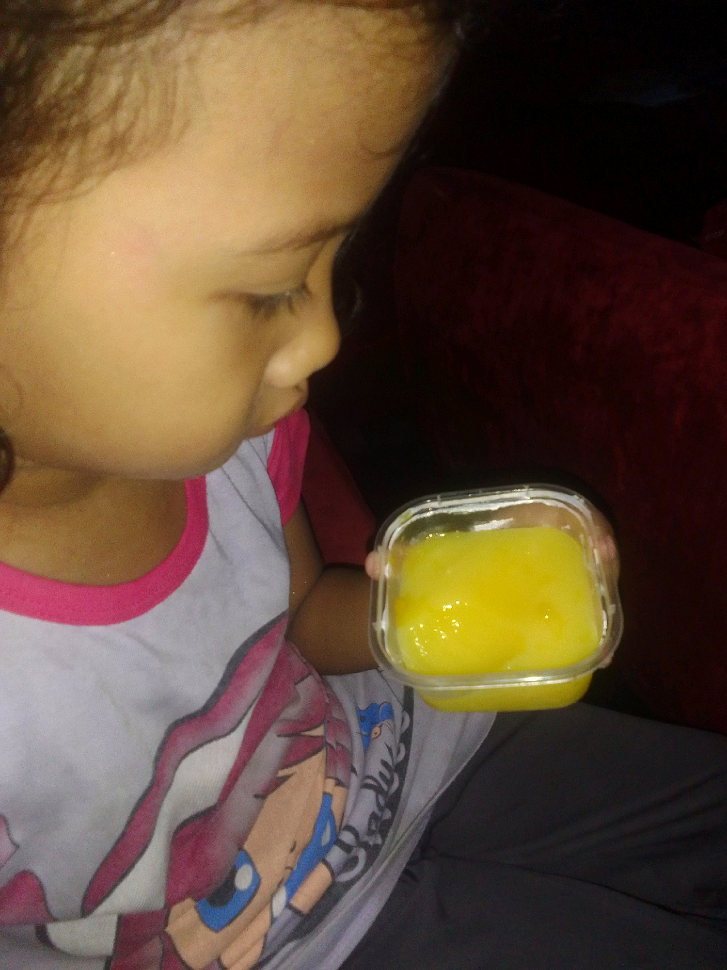 A close-up, profile shot shows a young child holding a small, clear, square plastic container filled with yellow pudding. The child's face is illuminated from the side, highlighting their cheek and eye. They are wearing a white t-shirt with a pink trim around the neck and a cartoon graphic. The child appears to be sitting on a dark-colored surface, possibly a sofa or chair, which is partially visible in the background. The setting is indoors and dimly lit, with the red upholstery of furniture visible behind the child. The image does not provide any specific geographical indicators that would suggest a location in Ngadiluwih, Indonesia.