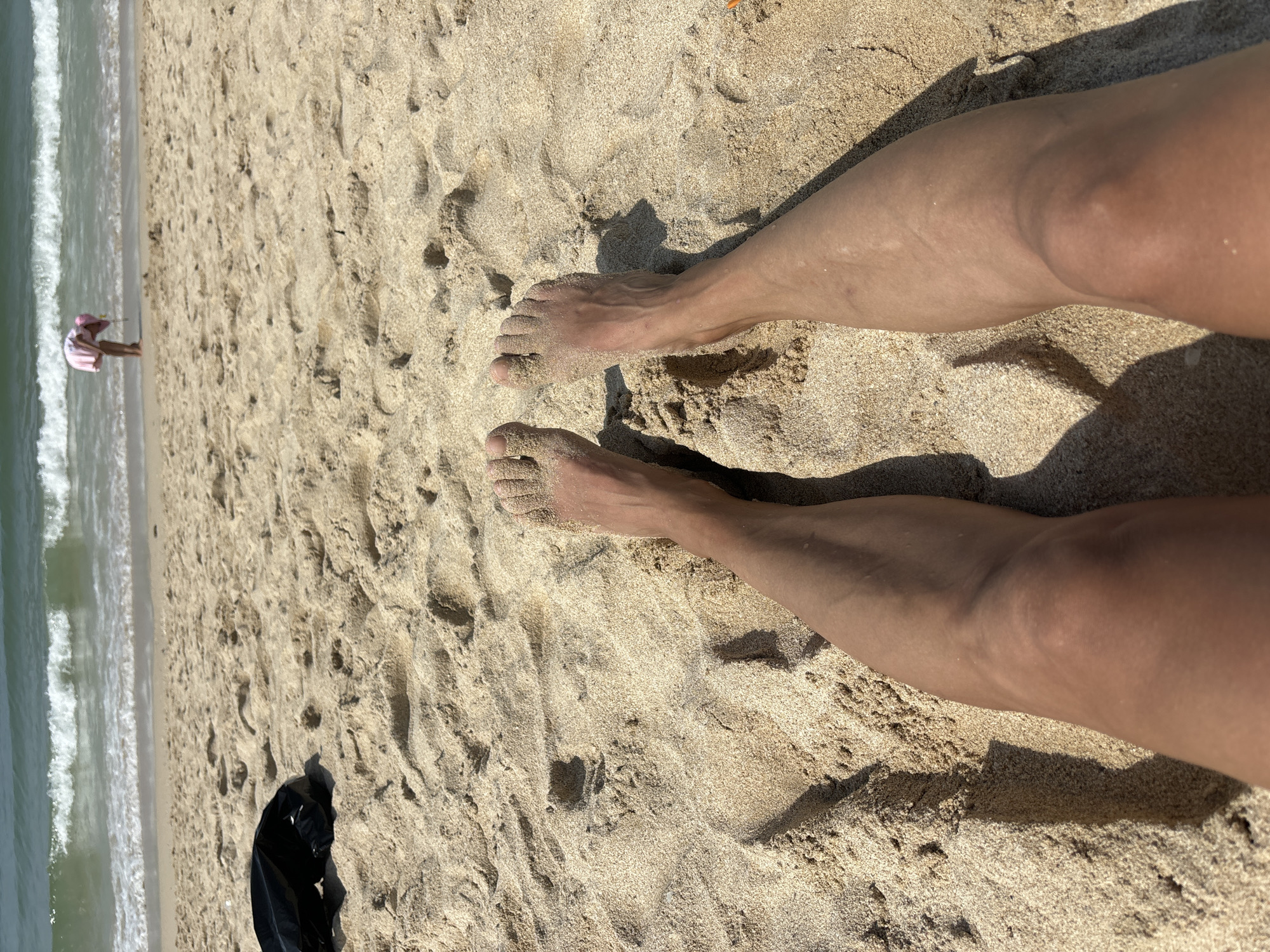 A close-up, high-angle view of two bare feet and lower legs covered in sand on a beach. The sand is light brown and textured with footprints and ripples. In the background, the ocean is visible with gentle waves and a shoreline. Further in the distance, a small person wearing a pink outfit and hat is bent over near the water. A black plastic bag is lying on the sand to the left of the feet. The scene suggests a sunny day at the beach.