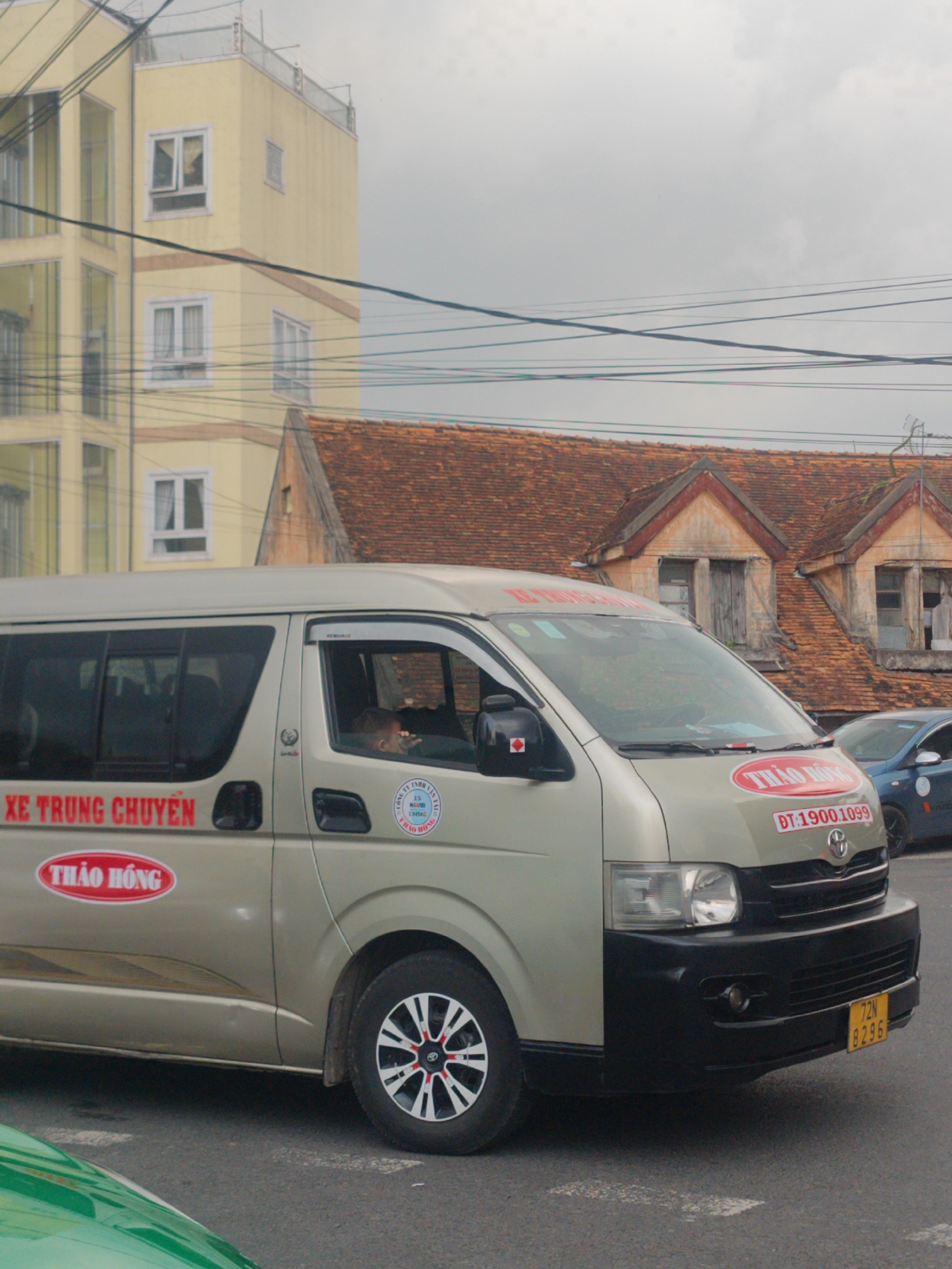 The image presents an urban street scene in Cam Ly - Đà Lạt, Vietnam, under an overcast sky. A prominent light beige Toyota Hiace van, serving as a shuttle bus, dominates the center of the frame. A driver is visible inside the vehicle. The van is clearly branded with "XE TRUNG CHUYỂN" (Shuttle Bus) and the company name "THẢO HỒNG," which appears in red text on multiple locations on the vehicle. A phone number, "ĐT: 1900.1099," is displayed on the front, and a license plate, "72N 8296," is visible on the bumper. A circular logo on the side includes "CÔNG TY TNHH" (Company Limited) and "THẢO HỒNG."
In the background, two distinct buildings illustrate a blend of architectural styles. To the left, a modern, light yellow multi-story building with numerous windows rises towards the sky. To the right, an older building with a distinctive red-tiled roof and two prominent dormer windows with wooden shutters adds a historical element to the setting. Overhead power lines crisscross the upper portion of the image. The presence of a partial green vehicle in the foreground and a blue car in the background indicates active street traffic. The scene captures a typical moment of daily life and transportation within a Vietnamese urban environment.