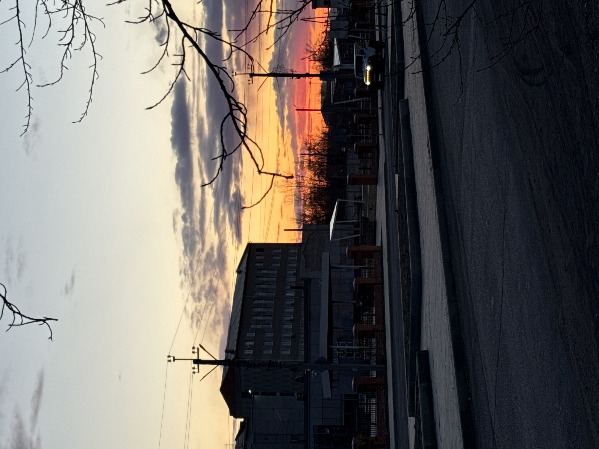 The image depicts a street scene in Borzya, Russia at sunset. A multi-story building with numerous windows dominates the lower portion of the frame. In the foreground, a paved street runs parallel to the building. Utility poles and wires are visible against the colorful sky, which displays a gradient of orange, pink, and purple hues. Silhouetted tree branches frame the upper left corner of the image. A vehicle with its headlights on is parked along the side of the road. The overall lighting suggests it is dusk.