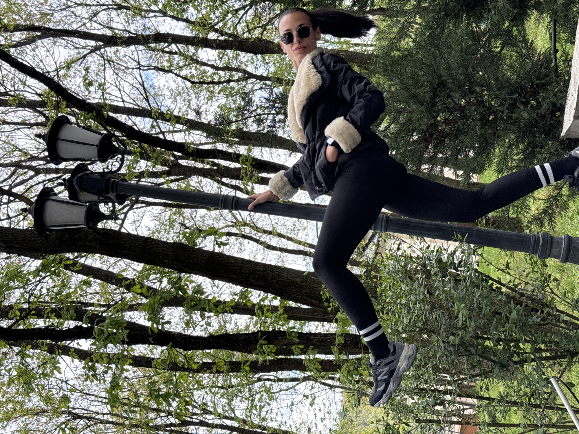 A woman strikes a dynamic, acrobatic pose in an outdoor setting, likely a park in Pleven, Bulgaria. She is positioned on the right side of the frame, extending her body and holding onto a black, ornate lamp post with her left hand. Her left leg is bent, and her right leg is extended backwards and upwards, suggesting a leap or a balancing act. She wears a black jacket with a cream-colored fluffy collar and matching cuffs, black leggings, and grey and black athletic sneakers with white socks that have two black stripes. Her dark hair is pulled back in a high ponytail, and she wears round sunglasses, looking confidently towards the camera.
The lamp post, featuring two lanterns, spans horizontally across the middle of the image. The background is dominated by the canopy of deciduous trees with nascent, light green leaves, indicating early spring. The sky overhead is bright and overcast, providing diffused lighting. To the lower right of the frame, a dense evergreen bush or tree adds depth to the scene. The overall impression is one of athleticism and playfulness captured in a public green space. There is no visible text in the image.