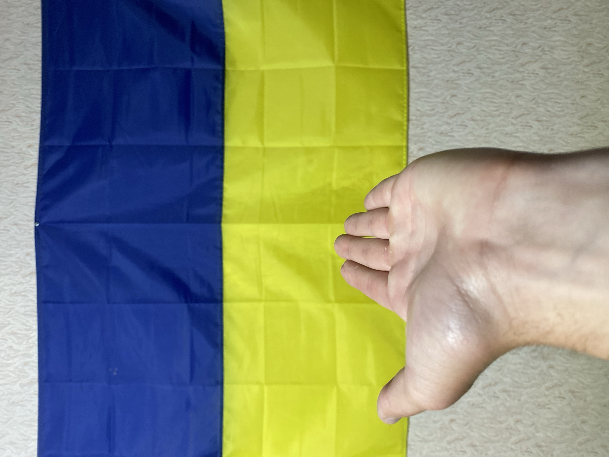 A photograph shows a close-up of a Ukrainian flag composed of horizontal bands of blue and yellow, with a hand partially obscuring the yellow portion of the flag. The background appears to be a textured, light-colored surface. The location in Poltava, Ukraine is not discernible from this image.