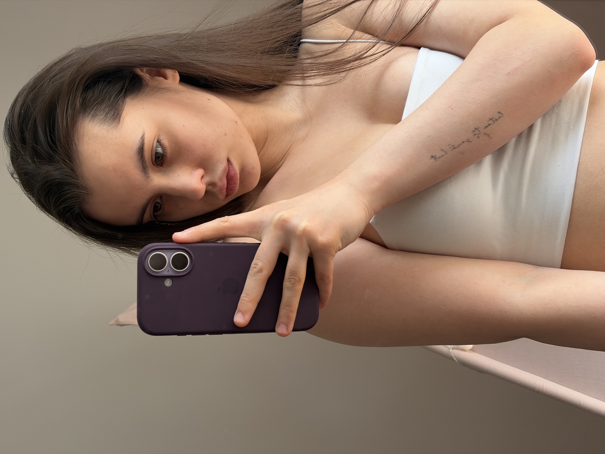 A young woman with long, dark brown hair and fair skin is captured in a close-up, rotated portrait. She appears to be lying down, holding a dark purple smartphone with both hands, positioned to take a selfie or interact with the device. Her head is tilted, and her gaze is directed towards the phone or slightly past it, with a calm, somewhat pensive expression.
She is wearing a white bralette or crop top with thin straps. A notable detail is a handwritten script tattoo on her inner right forearm. The text is mirrored in the image, and when unmirrored, it reads "Find time to travel." Her skin shows natural details like faint freckles on her face and some creasing on her neck, indicative of her pose. Her fingernails are short and unpolished.
The background is a plain, light, neutral-colored surface, suggesting an indoor setting. The lighting is soft and natural, indicating it is daytime. The image conveys a casual, intimate moment. This scene takes place in Chaikovsky, Russia.