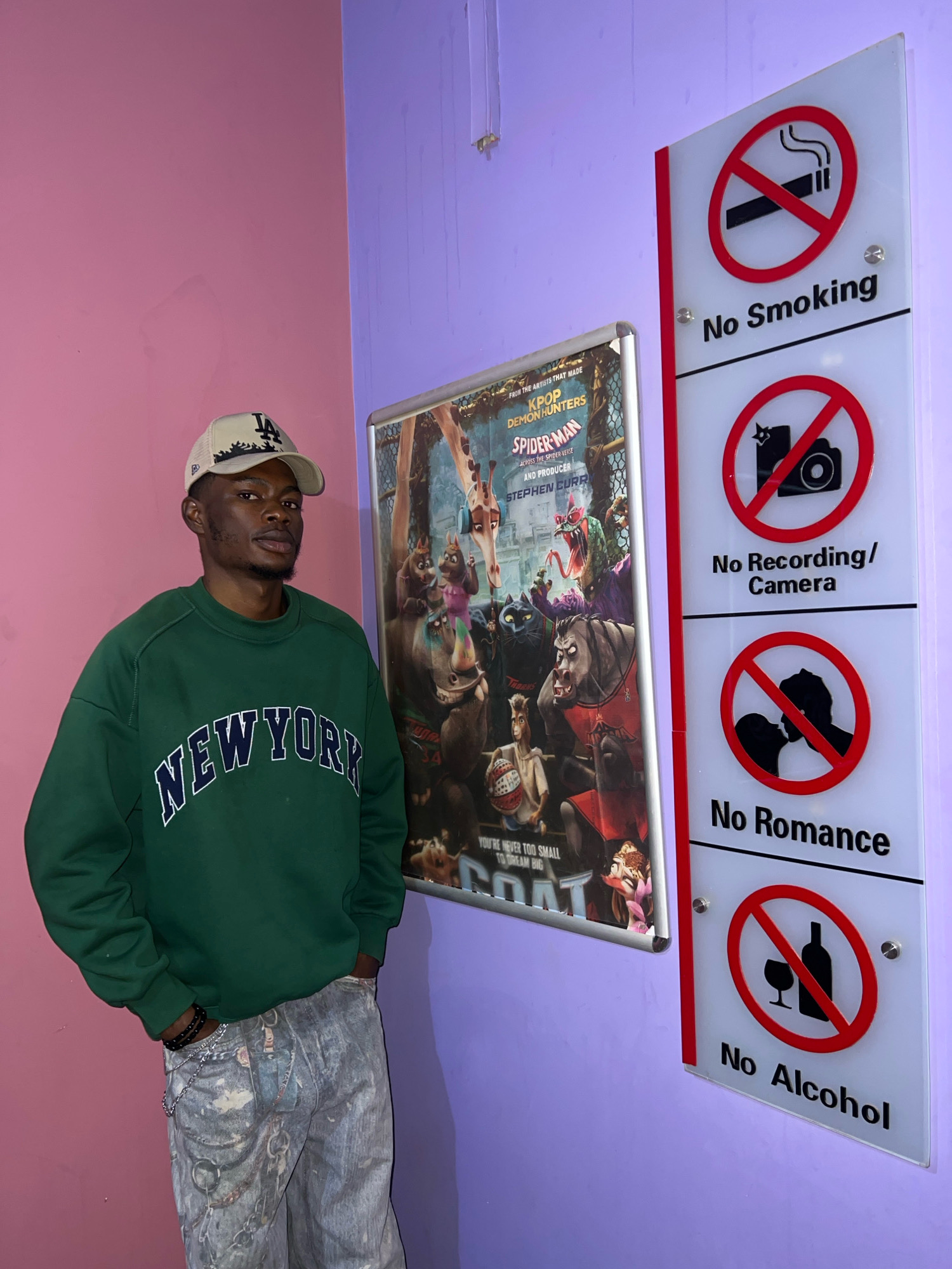 A young Black man stands against a pink and purple wall, posing for a photo. He wears a green sweatshirt with "New York" printed in white letters, a beige baseball cap, and patterned jeans. To his right, a sign displays four rules: "No Smoking," "No Recording/Camera," "No Romance," and "No Alcohol," each accompanied by a red circle with a diagonal line through it. A movie poster, featuring characters from "Spider-Man: Across the Spider-Verse," is also visible on the wall. The scene is likely indoors, given the artificial lighting and the presence of the wall and poster. No specific geographical landmarks of Jos, Nigeria, are depicted, therefore the location in Jos, Nigeria cannot be confirmed based on the image alone.