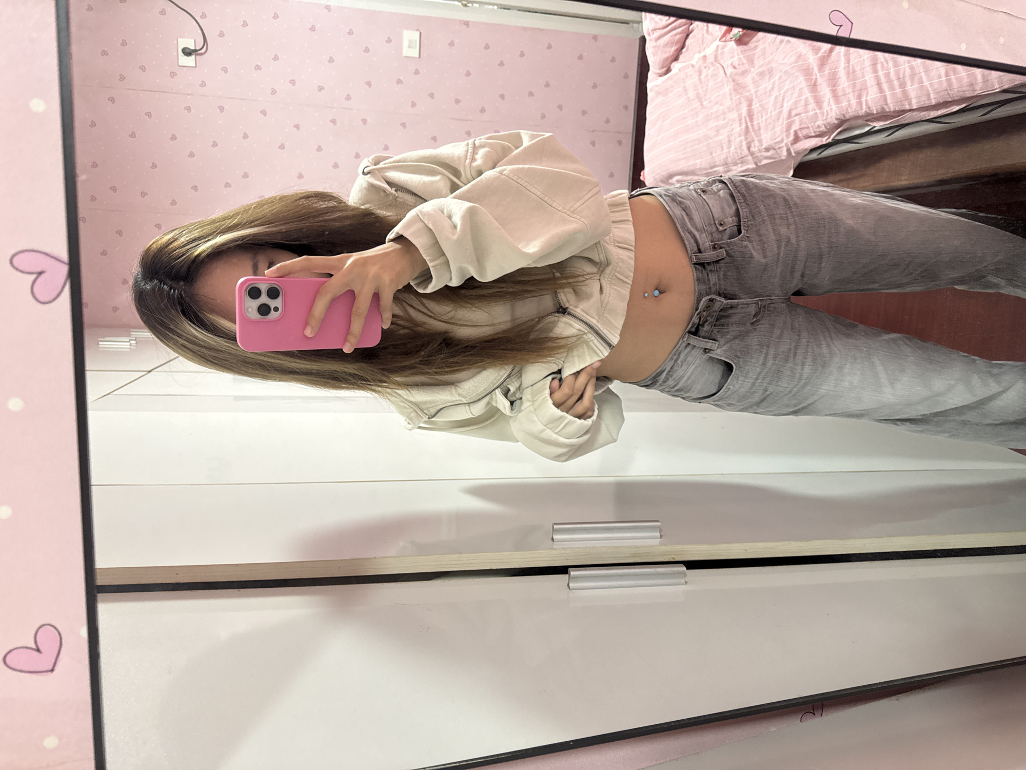 A young woman with long brown hair stands in front of a mirror taking a selfie with a pink smartphone. She is wearing a beige jacket that is slightly unzipped, revealing her midriff, and grey jeans. A navel piercing is visible. The background includes a pink wall with heart patterns, a bed with striped pink bedding, and a white wardrobe with silver handles. The scene appears to be indoors, possibly a bedroom. There is no visible evidence to indicate the location is in Shapingba District, China.