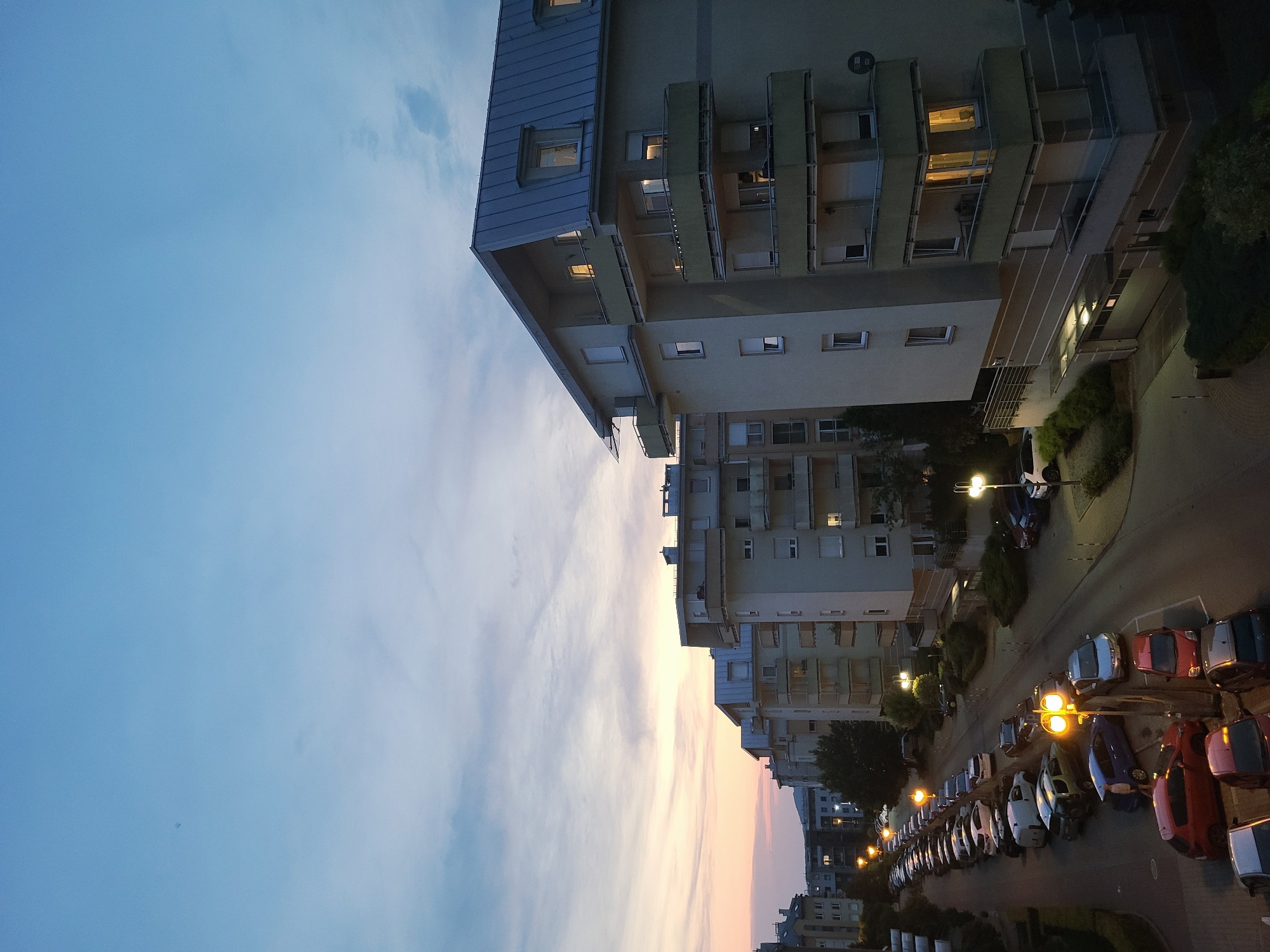 An eye-level, medium shot captures a street lined with parked cars and apartment buildings at dusk in an urban setting. The sky displays a gradient of blue, transitioning to soft hues of pink and orange near the horizon. The buildings, constructed from light-colored materials, feature multiple stories with balconies. Some windows are illuminated, indicating occupancy. A long row of parked vehicles, including sedans, SUVs, and vans, occupies the street. Streetlights cast a warm glow, illuminating the scene. Trees and shrubs are visible, adding a touch of greenery to the environment. The overall atmosphere suggests a peaceful evening in a residential area.