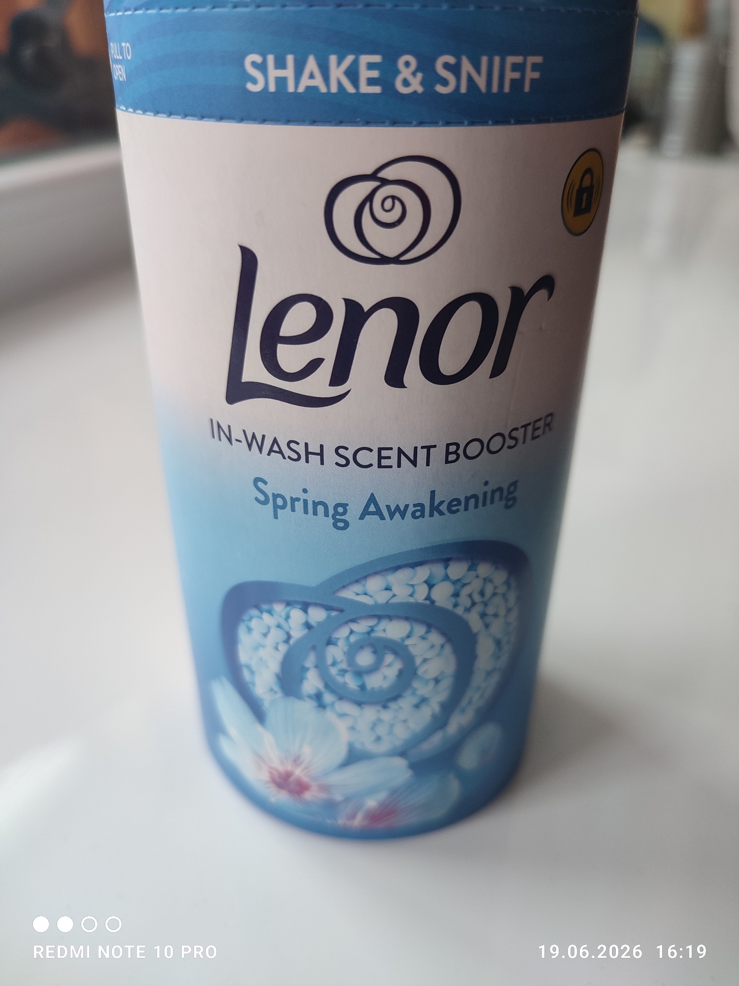 The image shows a close-up of a blue and white container of Lenor in-wash scent booster. The container has the text "SHAKE & SNIFF" at the top, followed by the Lenor logo and brand name. Below that, it reads "IN-WASH SCENT BOOSTER" and "Spring Awakening." The lower part of the container features a floral design with blue and white beads. In the bottom left corner, there is a watermark that says "REDMI NOTE 10 PRO," and in the bottom right corner, there is a timestamp that reads "19.06.2026 16:19." The background is blurred and appears to be a light-colored surface, possibly a table, with a hint of an orange object in the upper left corner. The scene is indoors, with no specific city or location details identifiable.