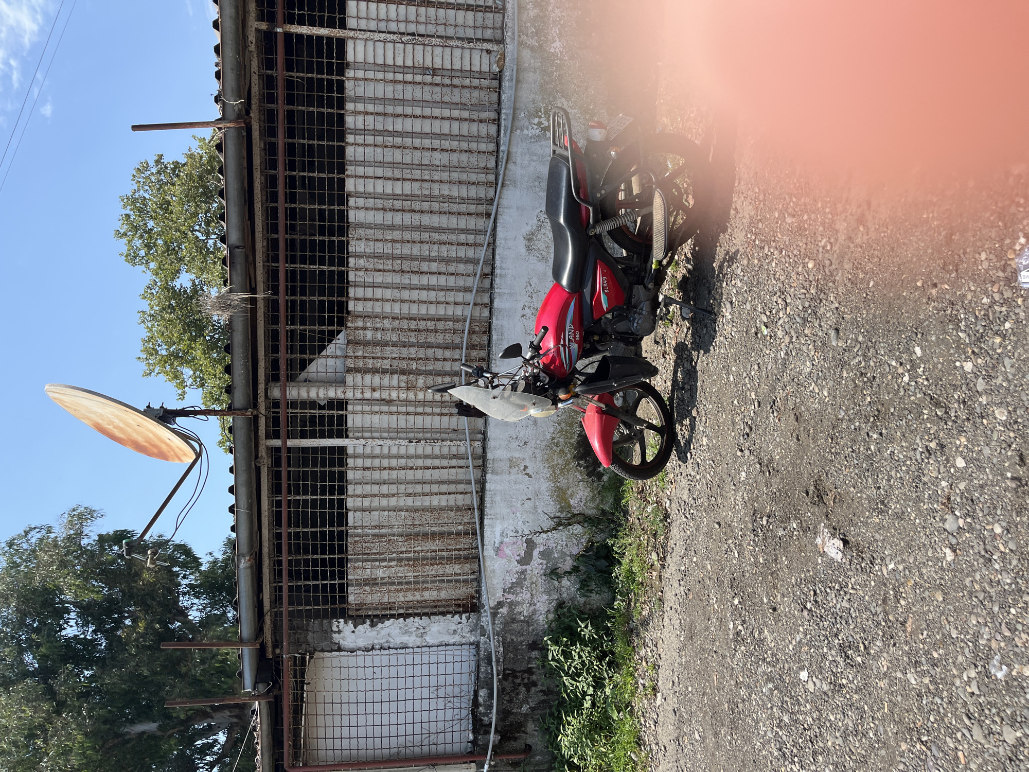 A red motorcycle is parked on a gravel surface in front of a weathered building. The building has a corrugated metal roof, a rusty metal grate covering its windows, and peeling paint on its lower section. A satellite dish is mounted on the roof. Green foliage grows along the base of the building. The sky is blue with a few white clouds. The scene appears to be outdoors, possibly in a rural or semi-urban area of Ureki, Georgia.