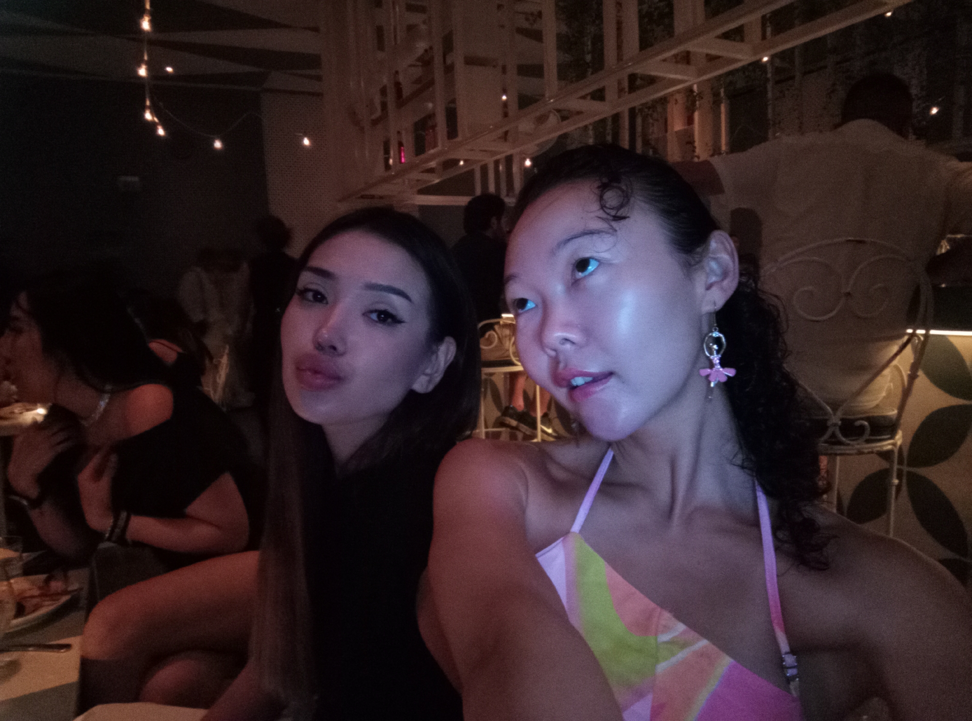 Two women are posing for a selfie in a dimly lit indoor setting, likely a restaurant or bar. The woman on the left has dark hair and is wearing a black top. The woman on the right has curly dark hair, light skin, and is wearing a colorful halter top. She is also wearing a pink earring. In the background, there are other patrons, lighting fixtures, and decorative elements. The location is possibly in North Kuta, Indonesia.
