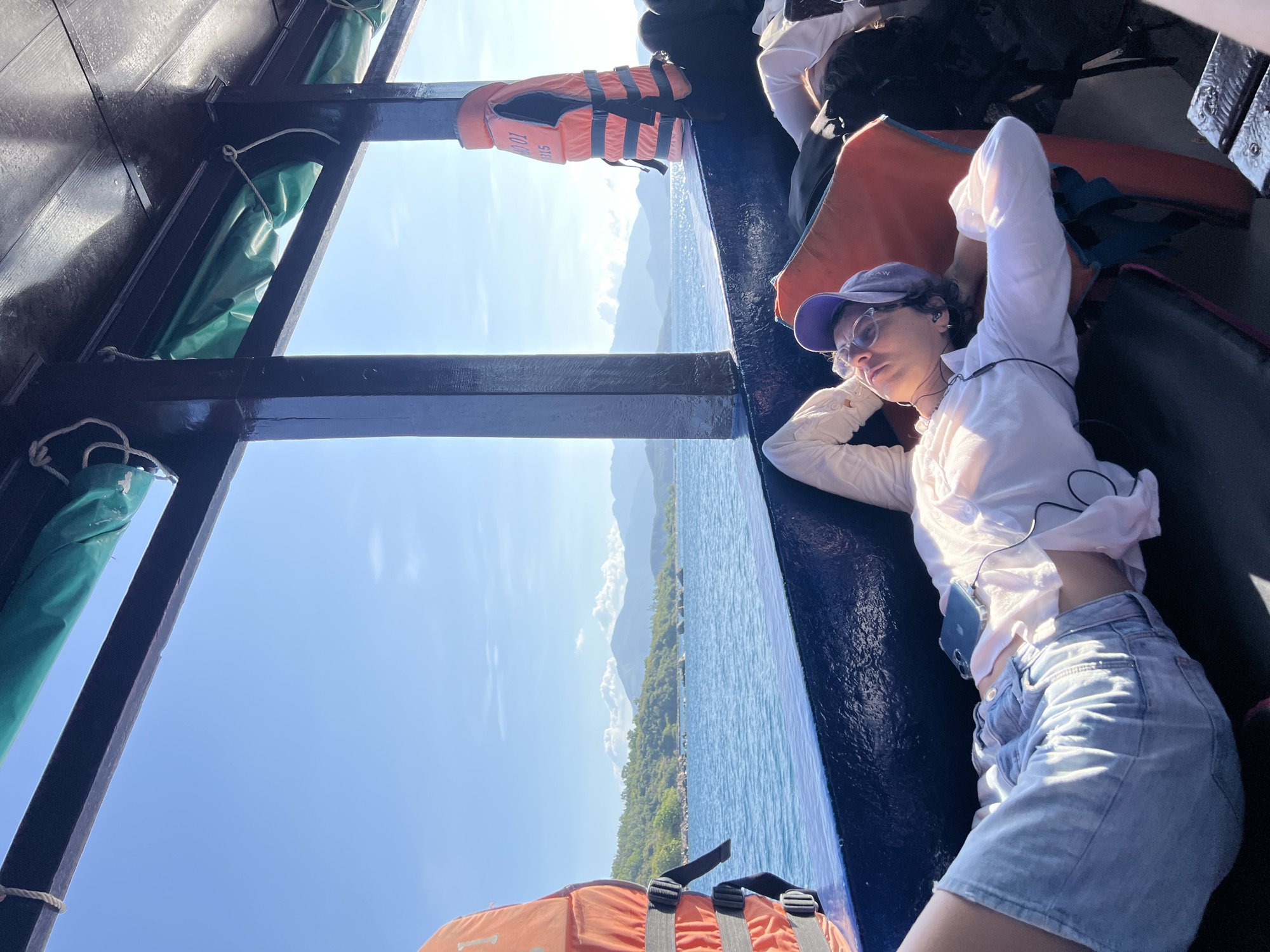 A young woman with short curly hair wearing a purple cap, sunglasses, a white long-sleeved shirt, and blue denim shorts is lying on a dark blue surface inside a boat. Her eyes are open, and she has earbuds connected to a phone resting on her stomach. Life vests are stored nearby. Through the open side of the boat, there is a view of a blue sea, a coastline with green vegetation, and mountains in the distance under a bright blue sky with some white clouds. The boat's dark wooden frame is visible. The image appears to be taken on a sunny day. The location appears to be within Nha Trang Ward, Vietnam, due to the coastal and mountainous landscape.